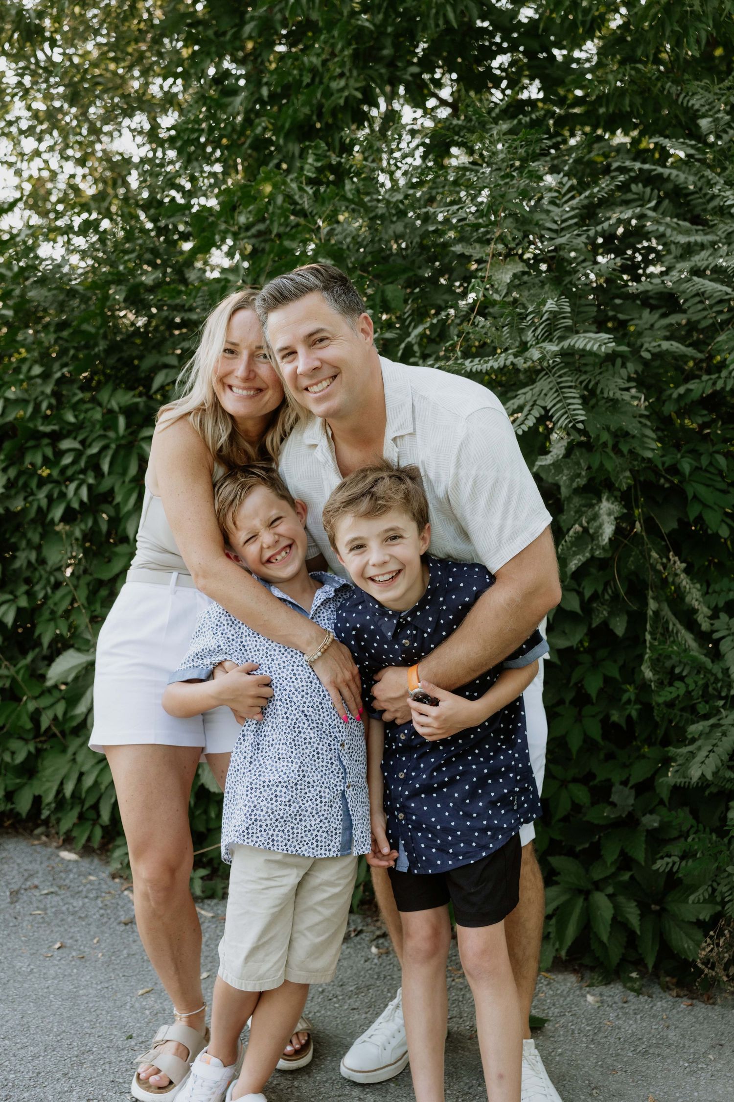 Happy family of four posing outdoors in front of lush green foliage, parents hugging two young boys.