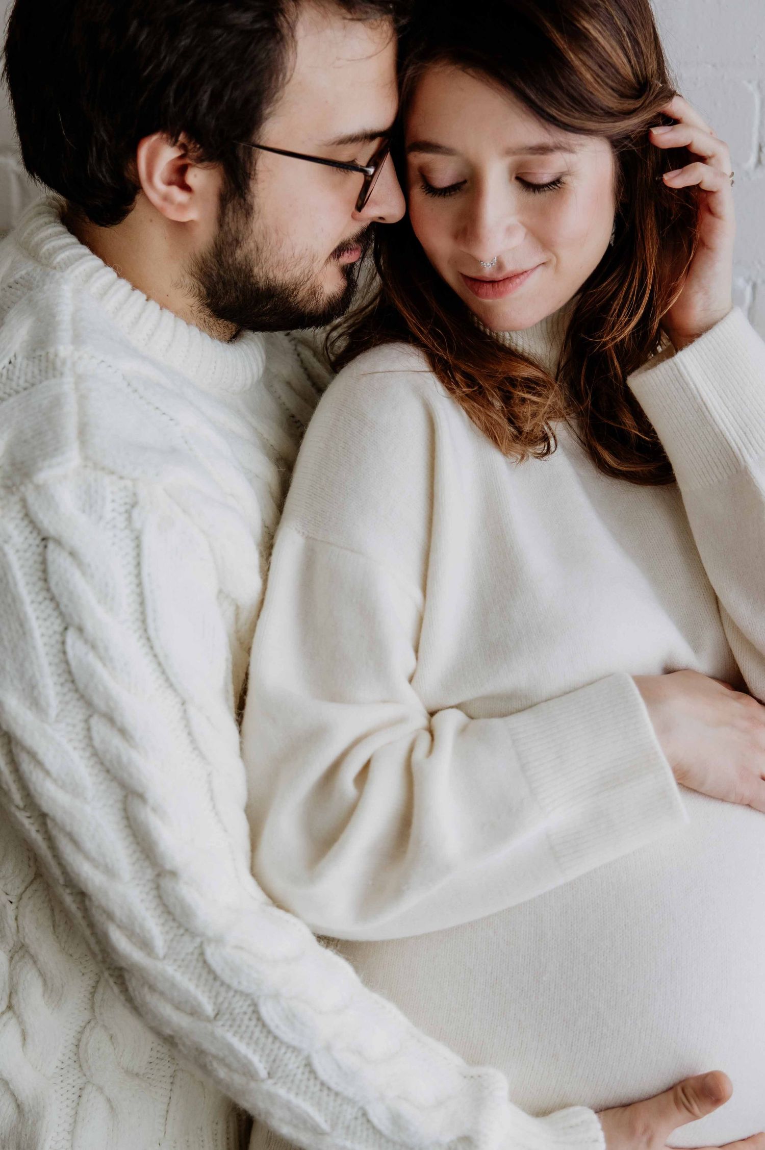 Intimate maternity photo of couple in white sweaters sharing tender moment.