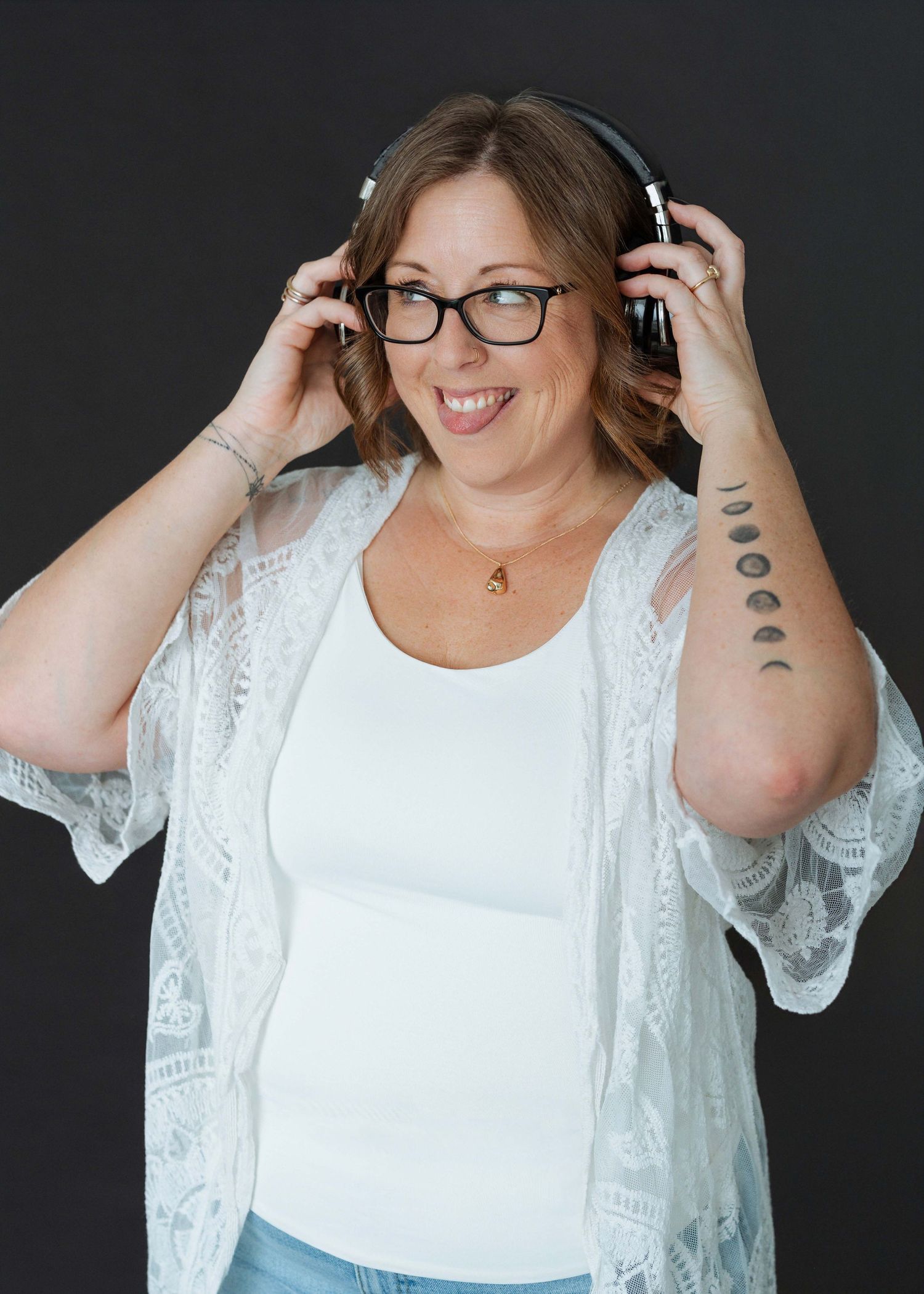 Smiling woman with glasses wearing headphones and a white lace cardigan, showing moon phase tattoo on arm.