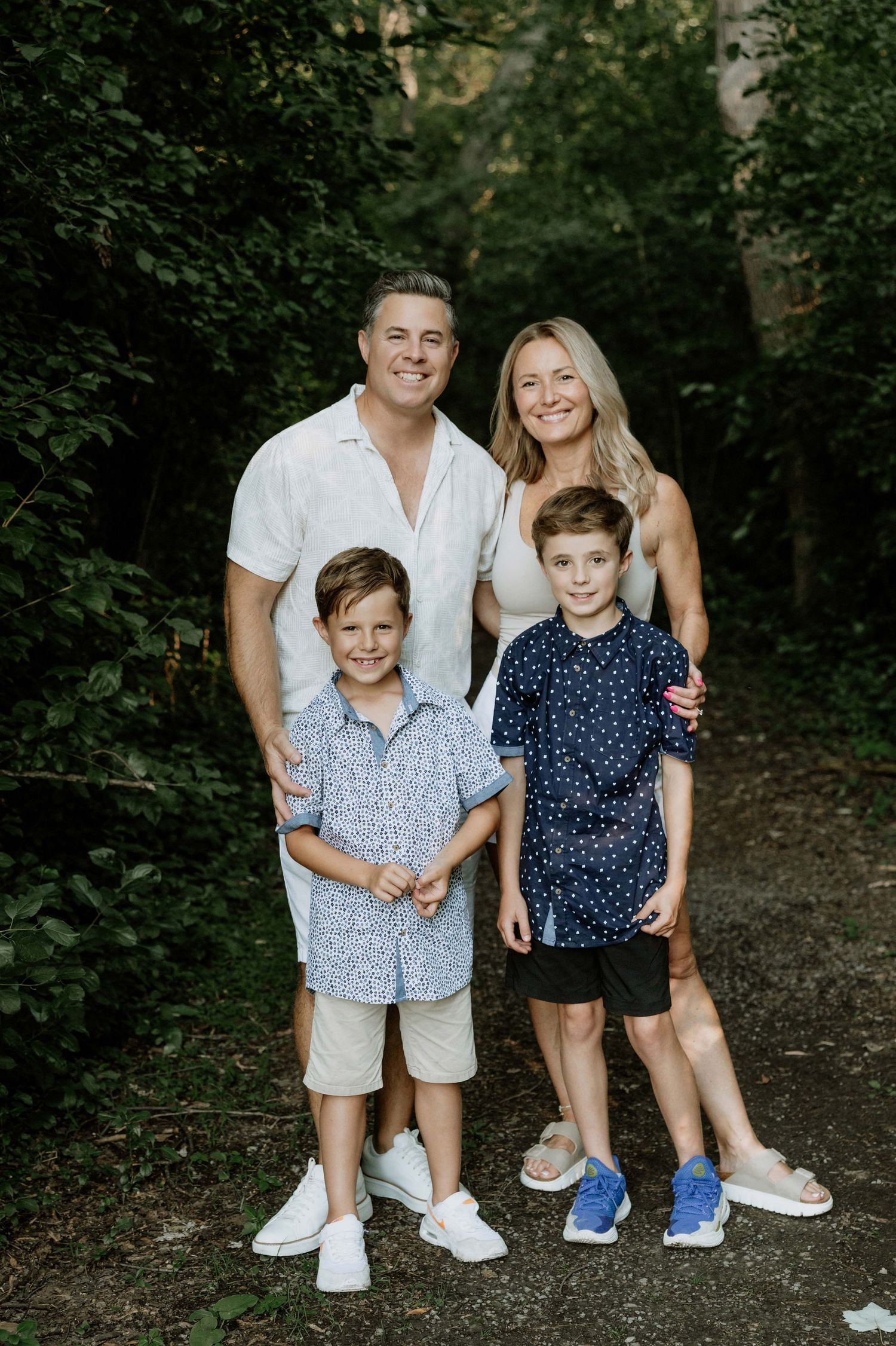 A smiling family of four poses outdoors on a wooded path, with two adults and two young boys dressed in casual summer attire.
