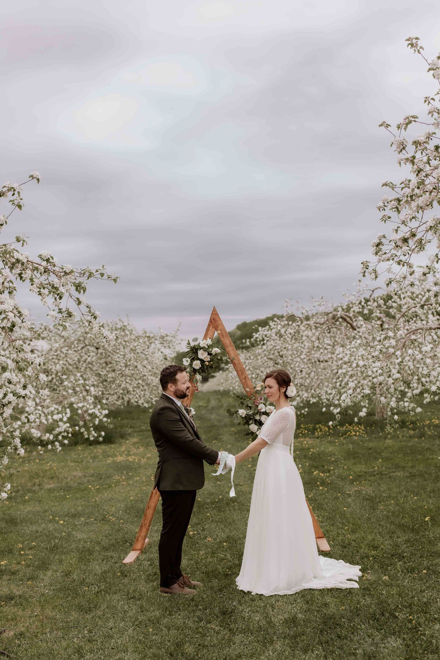Wedding couple poses playfully in blooming apple orchard under overcast spring sky.