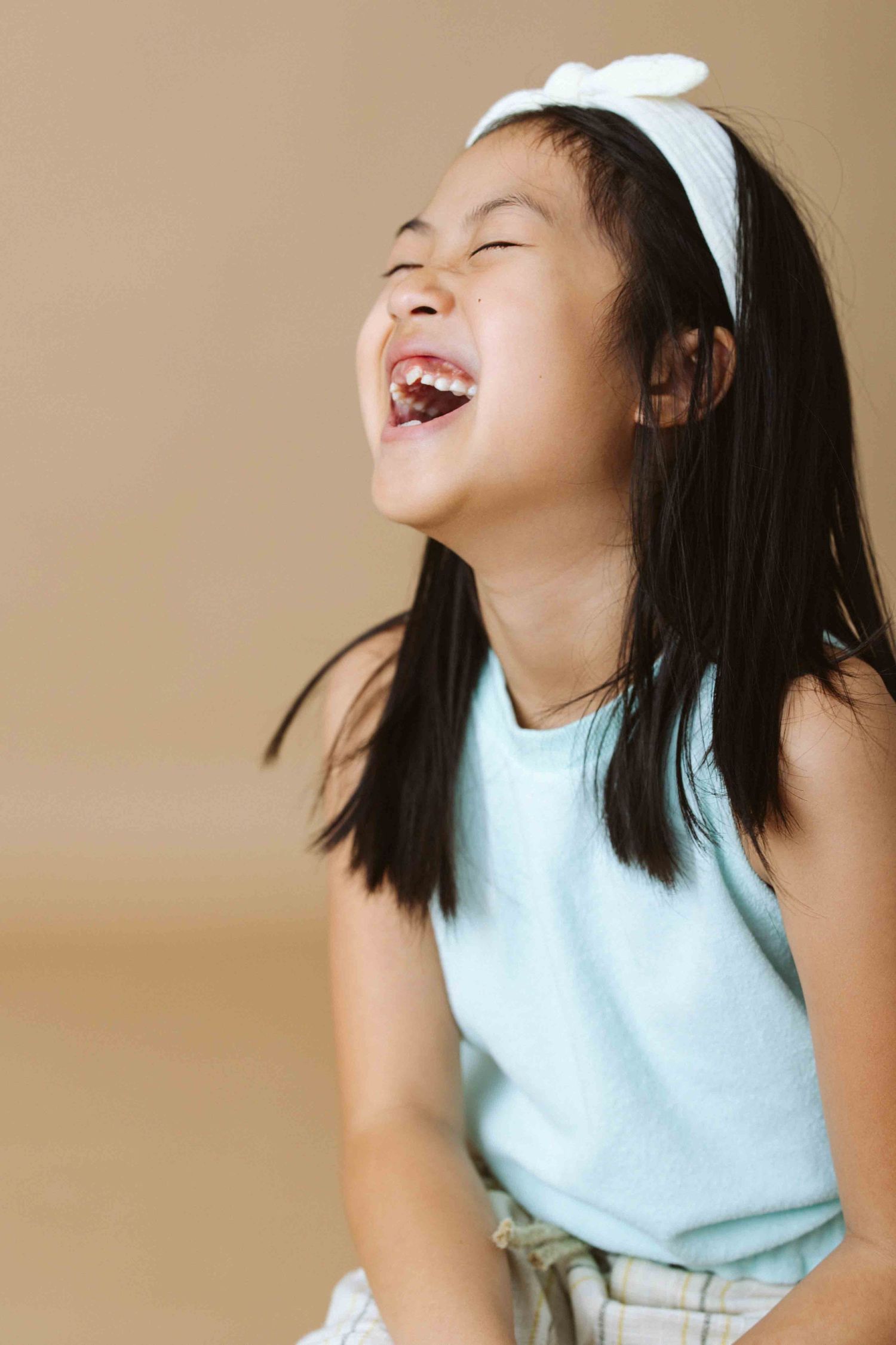 Young person in light blue top and white bow laughing joyfully against beige background.