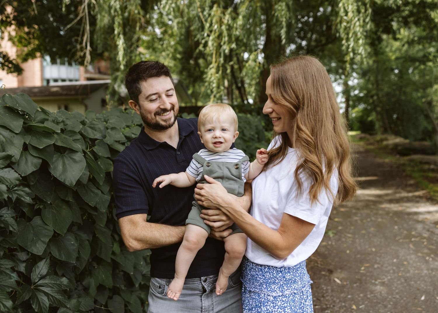 Happy young family with father holding baby boy smiling outdoors near lush green ivy wall.