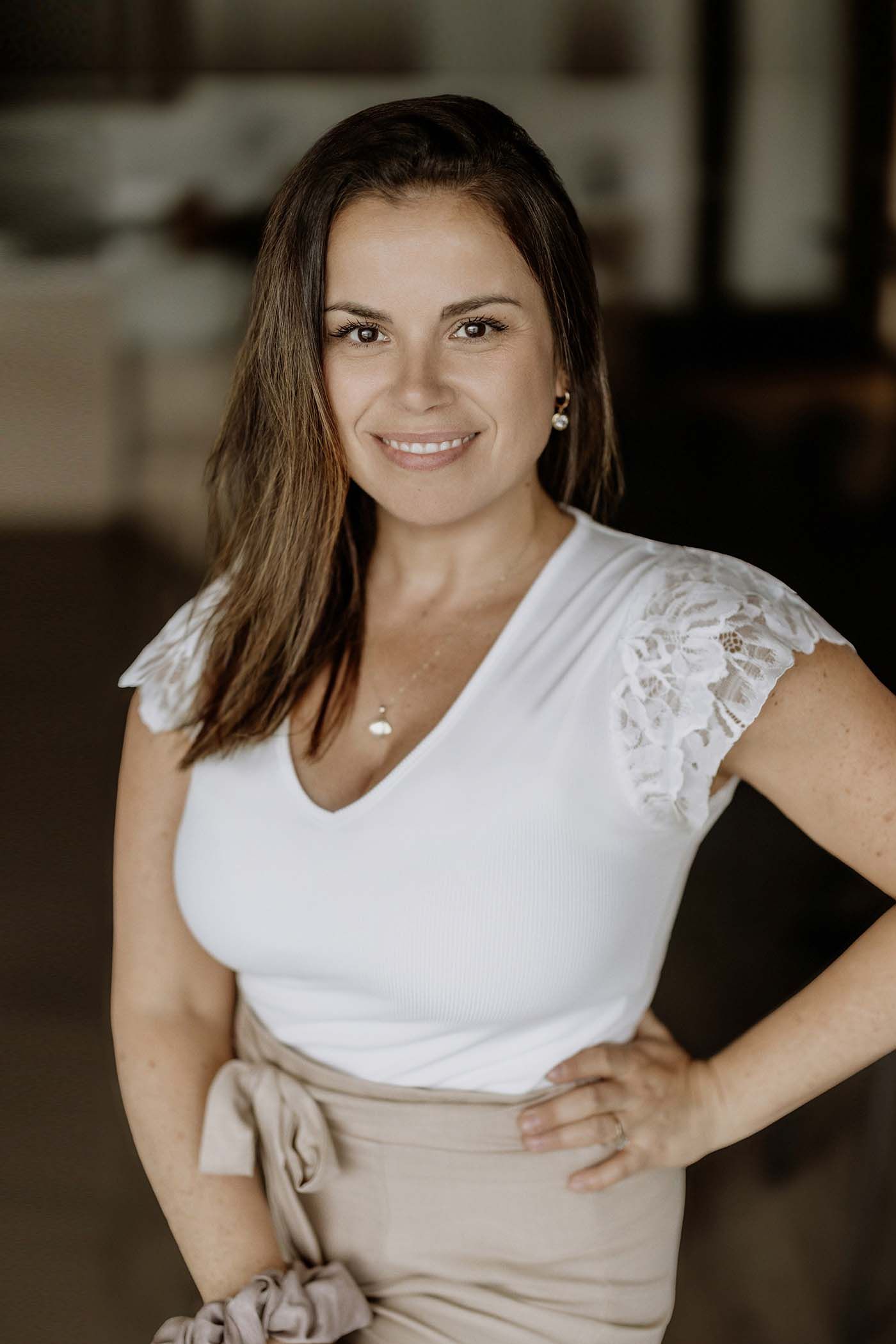 Smiling brunette woman in white lace top and beige skirt poses confidently with hand on hip.