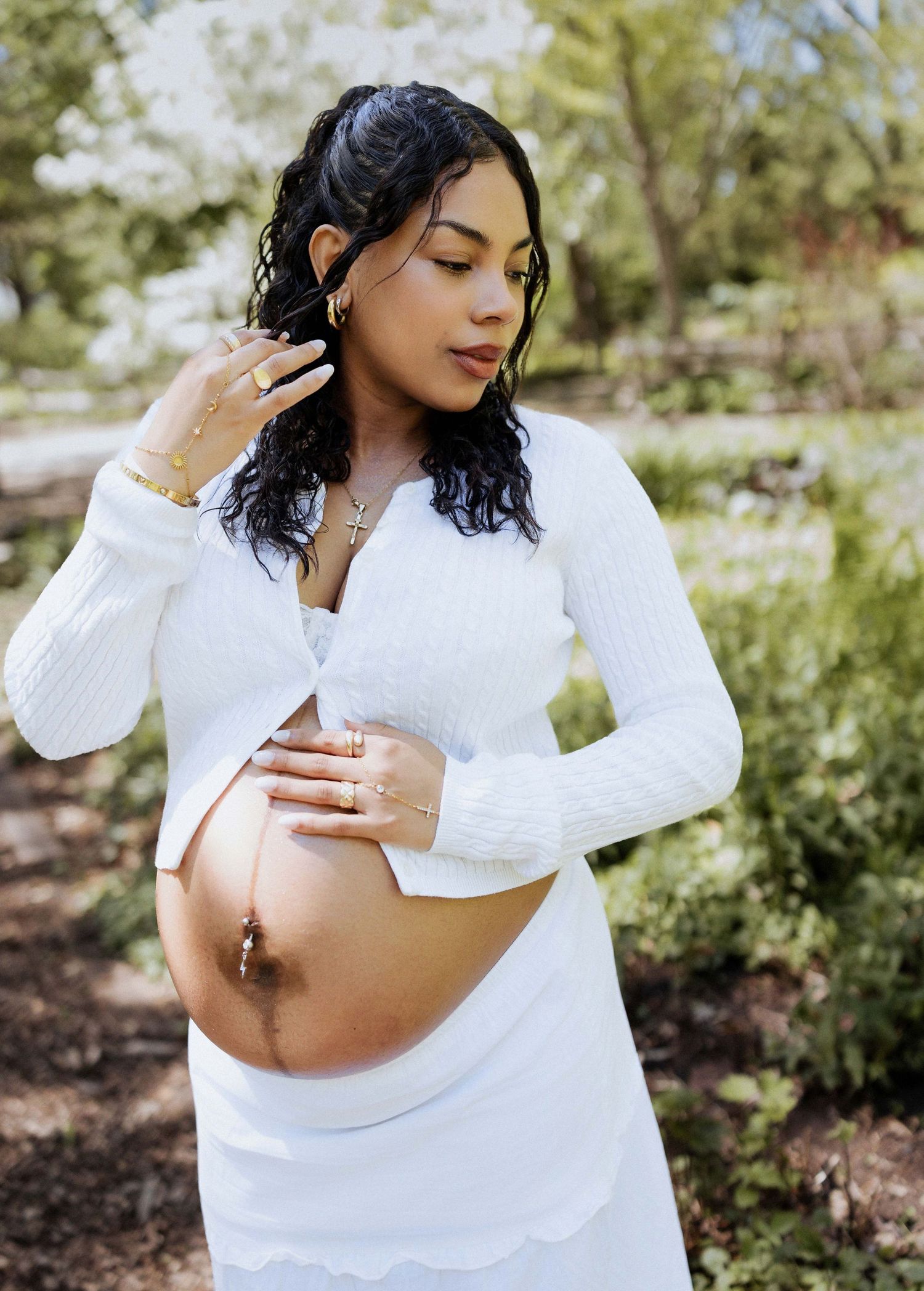 Pregnant woman in white crop top and skirt cradling baby bump outdoors, wearing gold jewelry.
