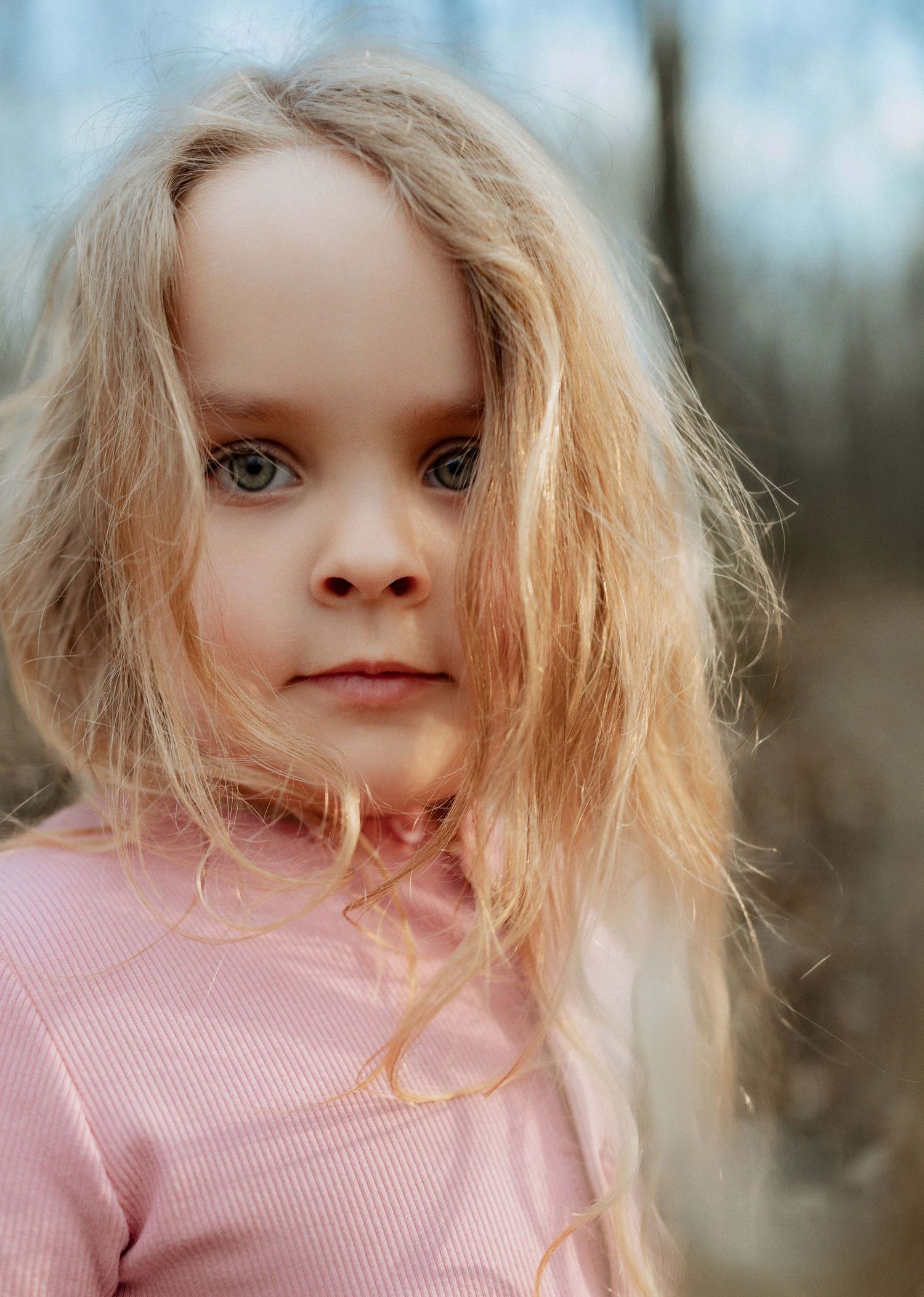 A young girl with long blonde hair and blue eyes wearing a pink shirt, photographed outdoors in soft natural light.