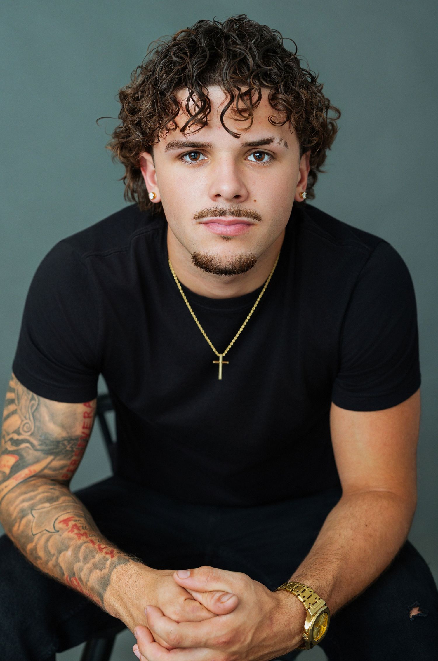 Young man with curly hair, tattoo sleeve, gold cross necklace and watch, wearing black t-shirt, studio portrait.