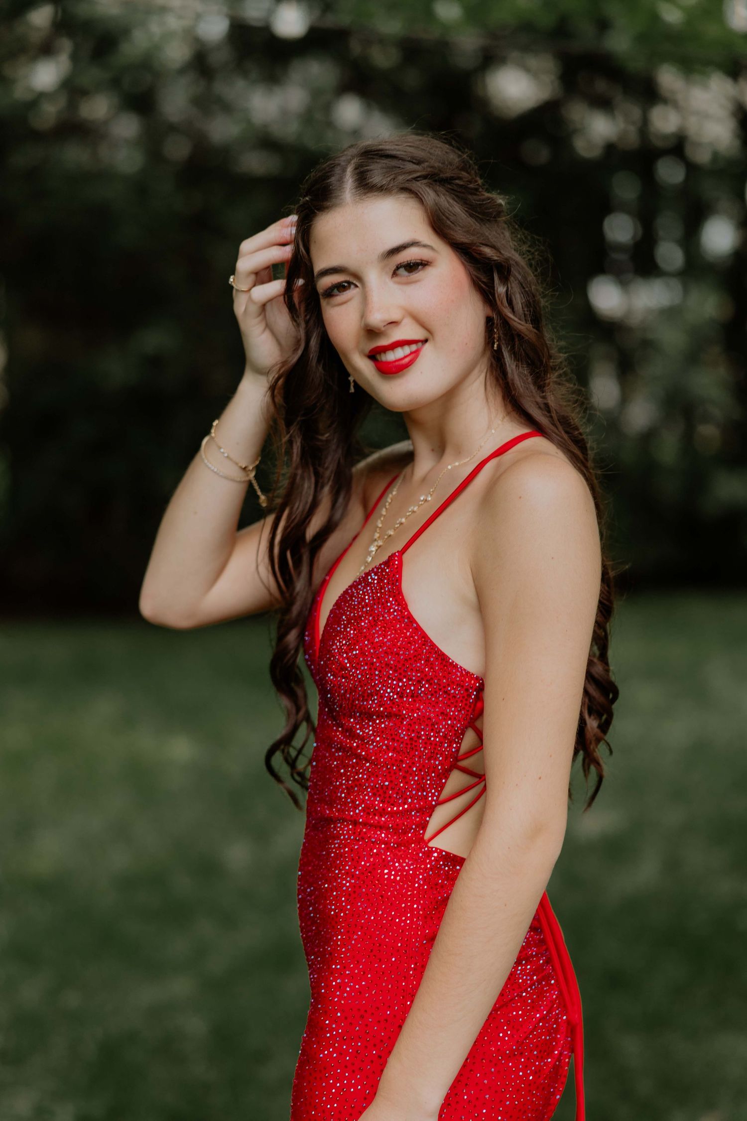 Young woman in a sparkling red sequin gown with matching lipstick, posing outdoors with lush green trees behind her.