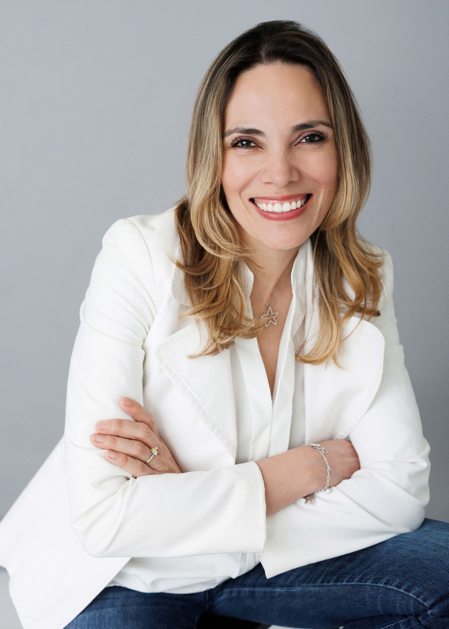 Professional portrait of someone in a white blazer and jeans against a gray backdrop.