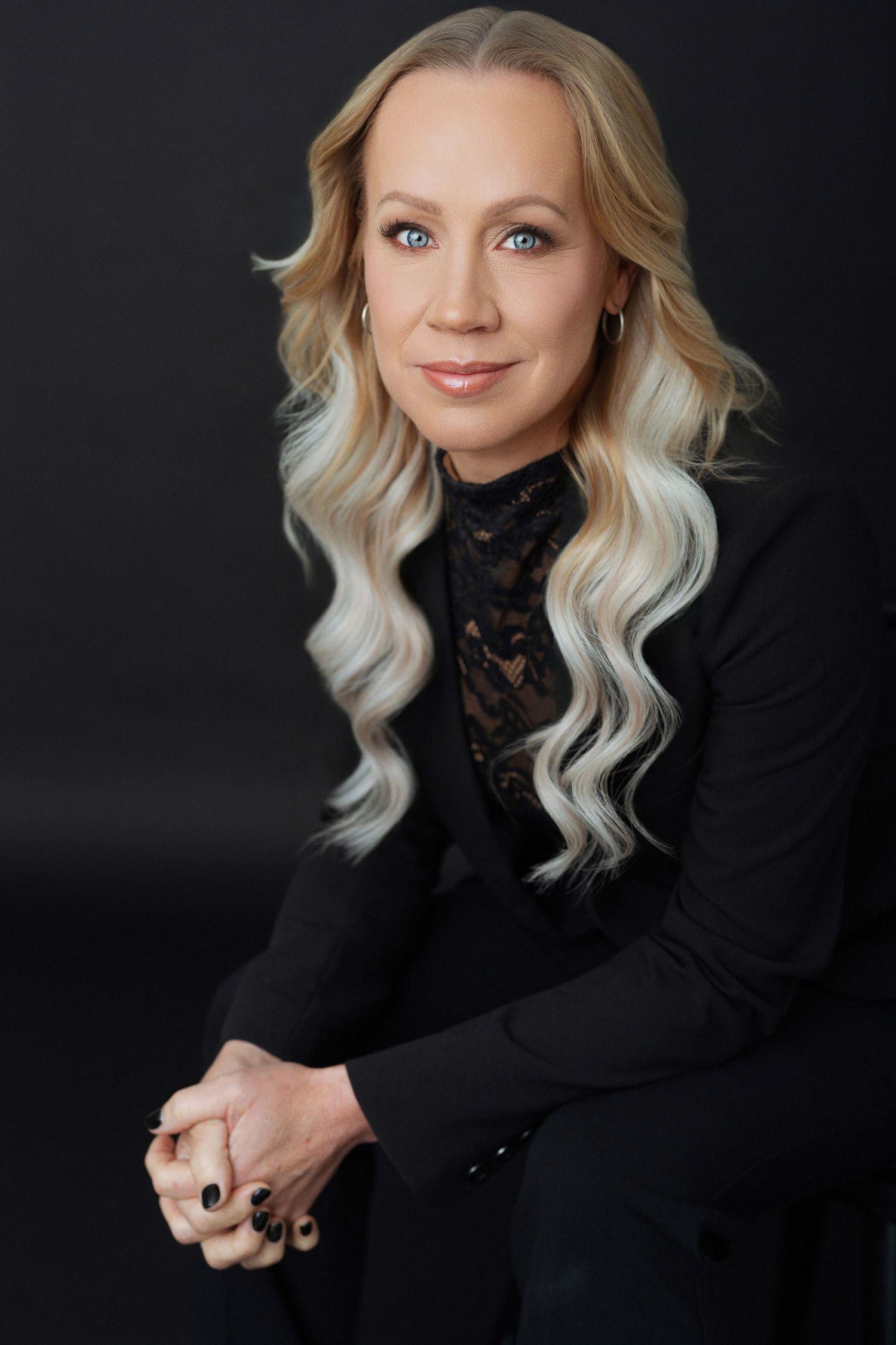 Professional woman with blonde wavy hair wearing a black blazer and lace top, posing confidently against dark background.