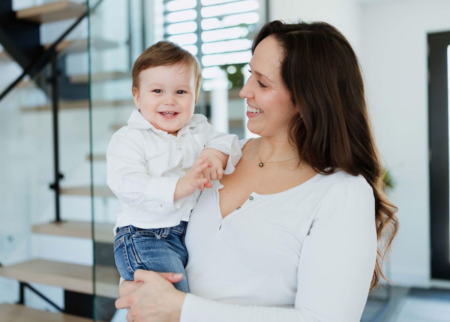 A smiling mother holds her happy baby boy in a modern home interior setting.