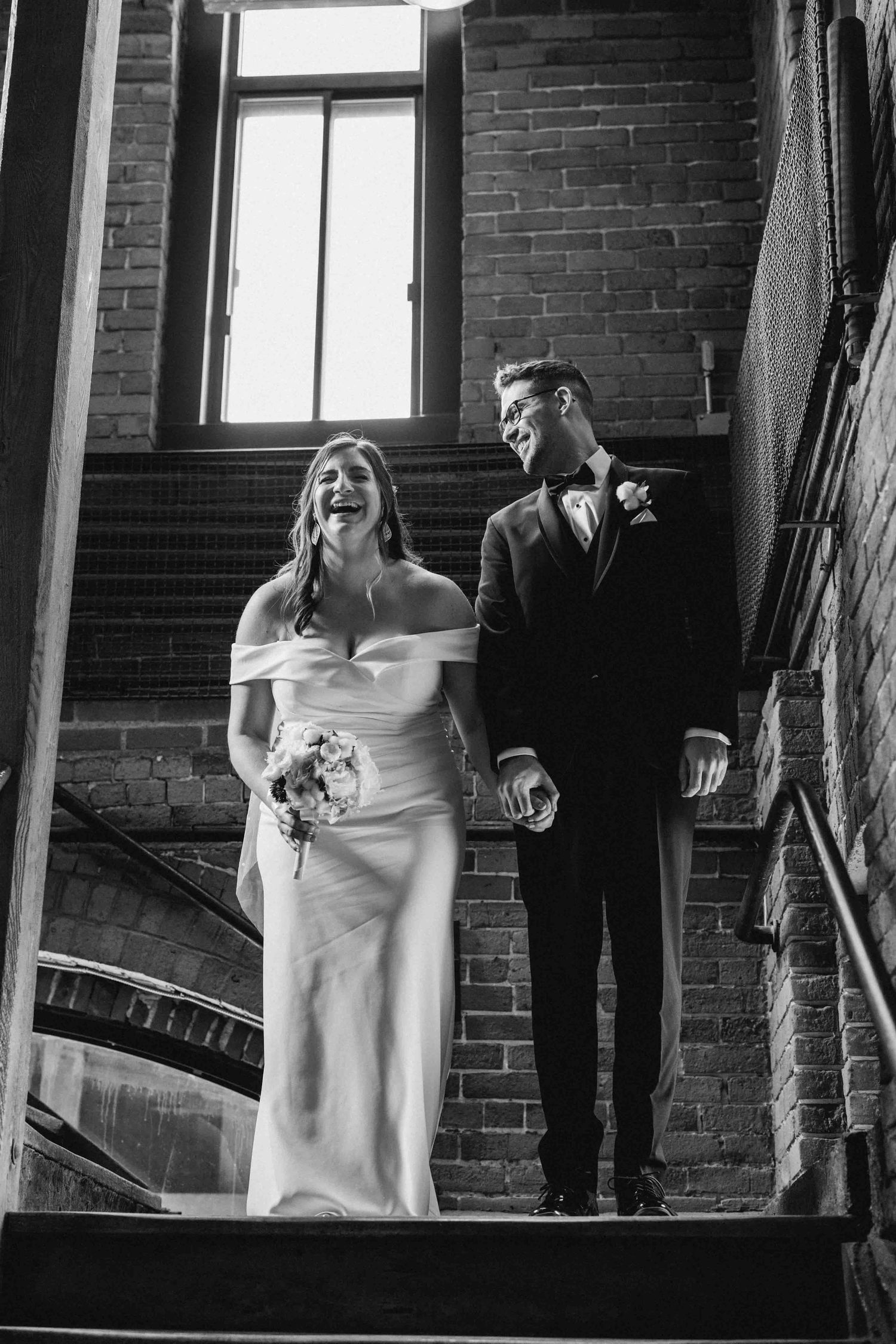 Black and white wedding photo showing bride and groom in dramatic lighting against exposed brick wall interior.
