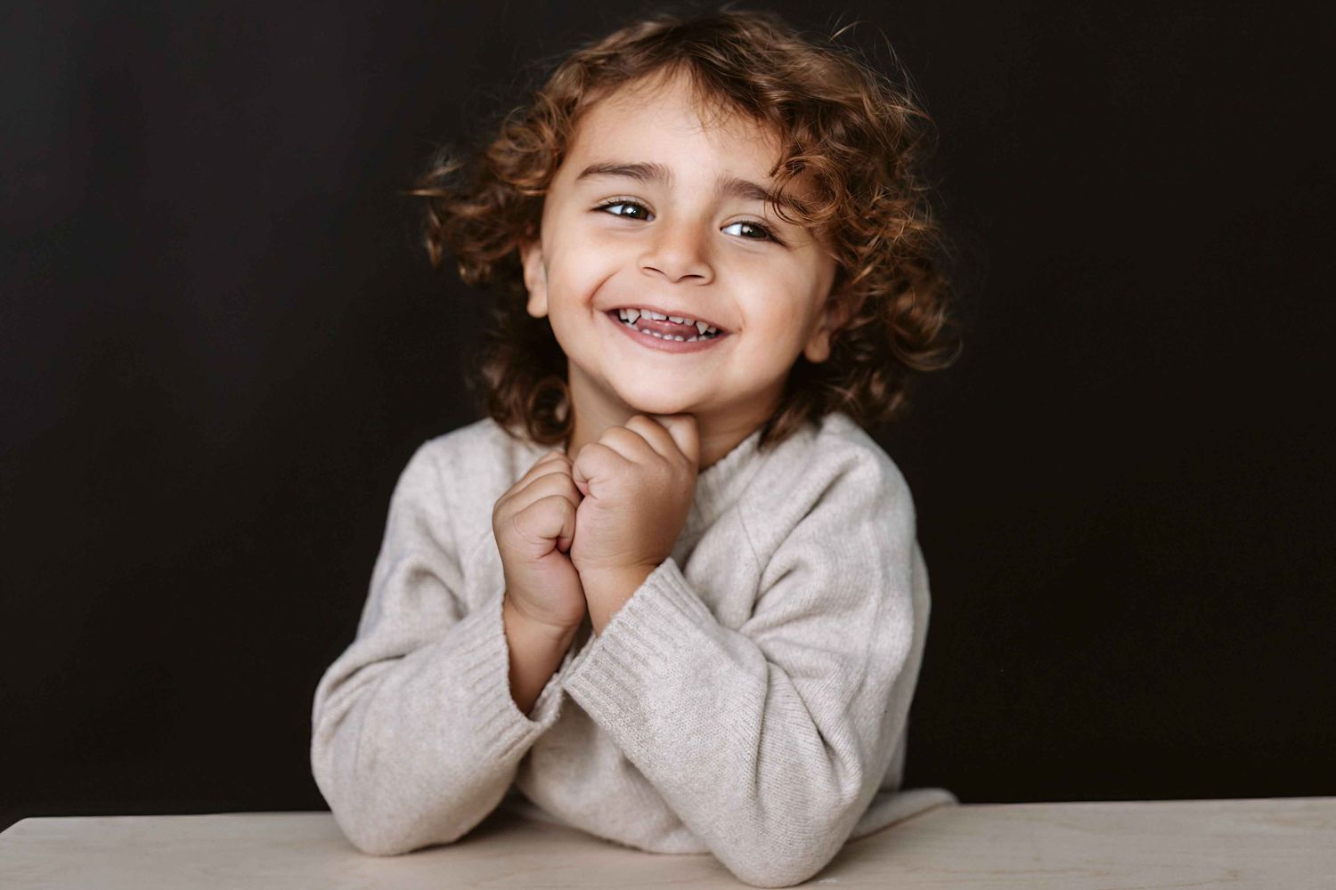 Child in light colored sweater poses playfully against dark background in portrait series.