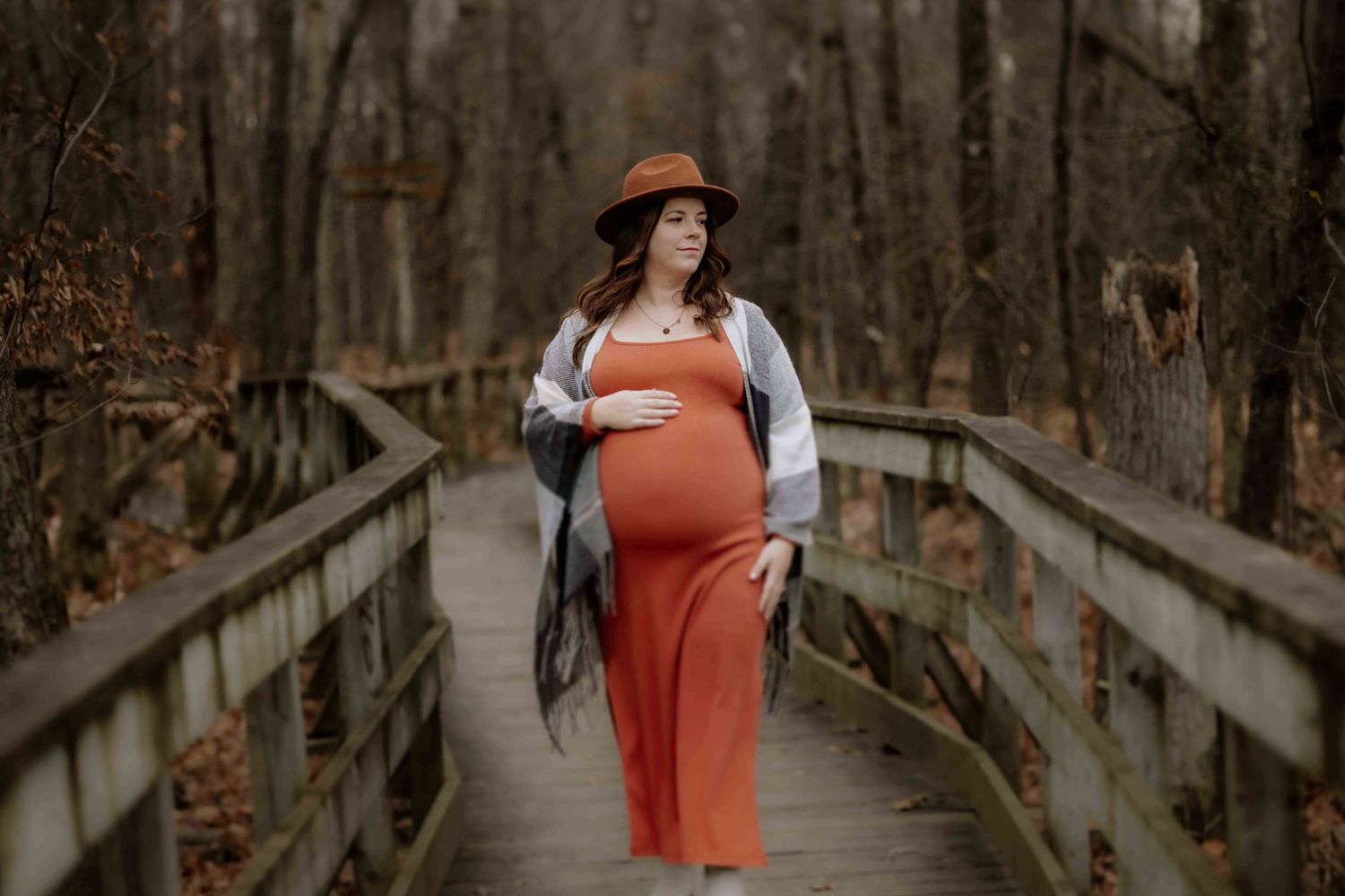 Pregnant woman in red dress and cardigan stands on wooden boardwalk in forest.