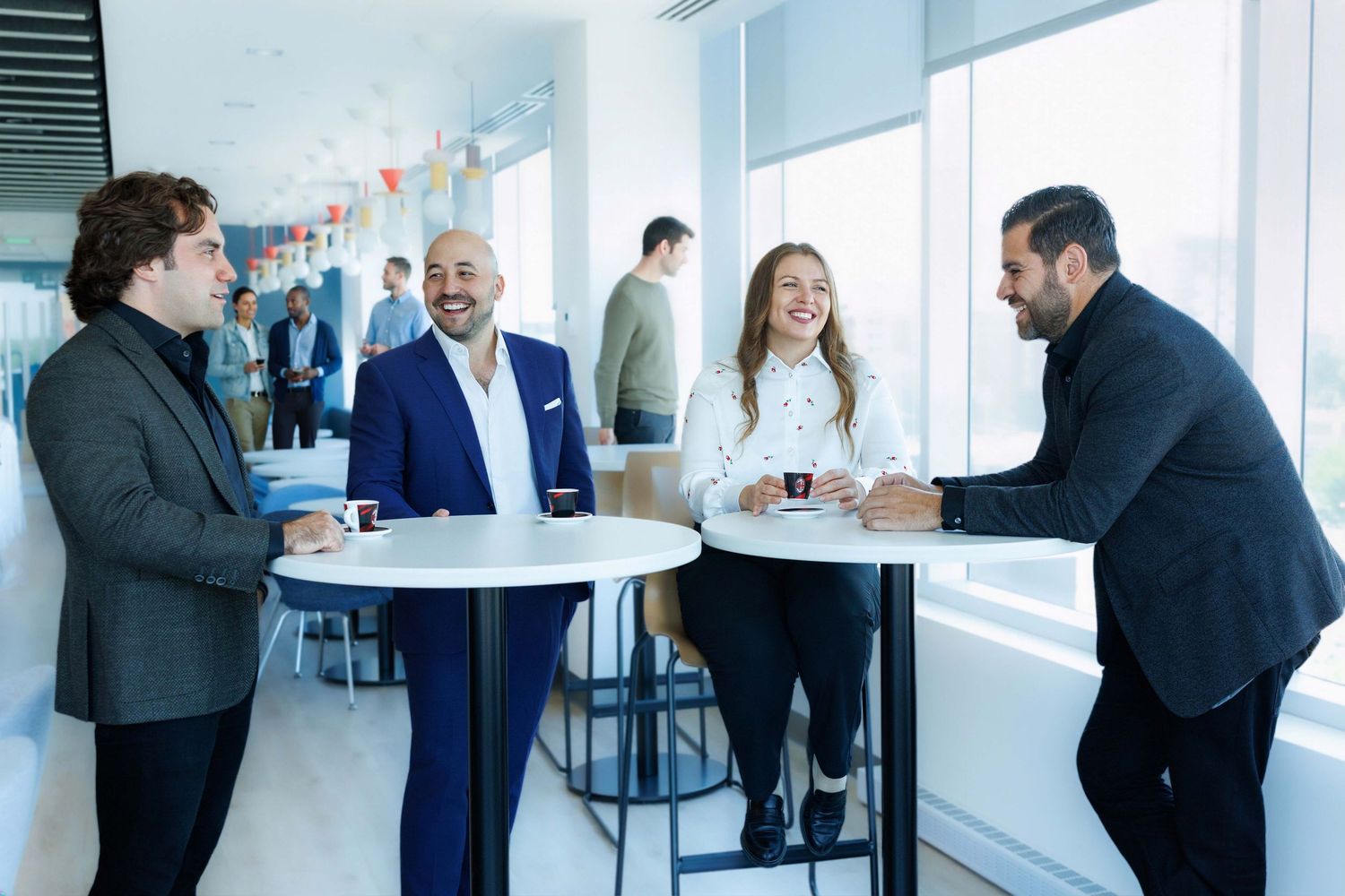 Four business professionals laughing and networking around a high-top table in a modern bright office space.