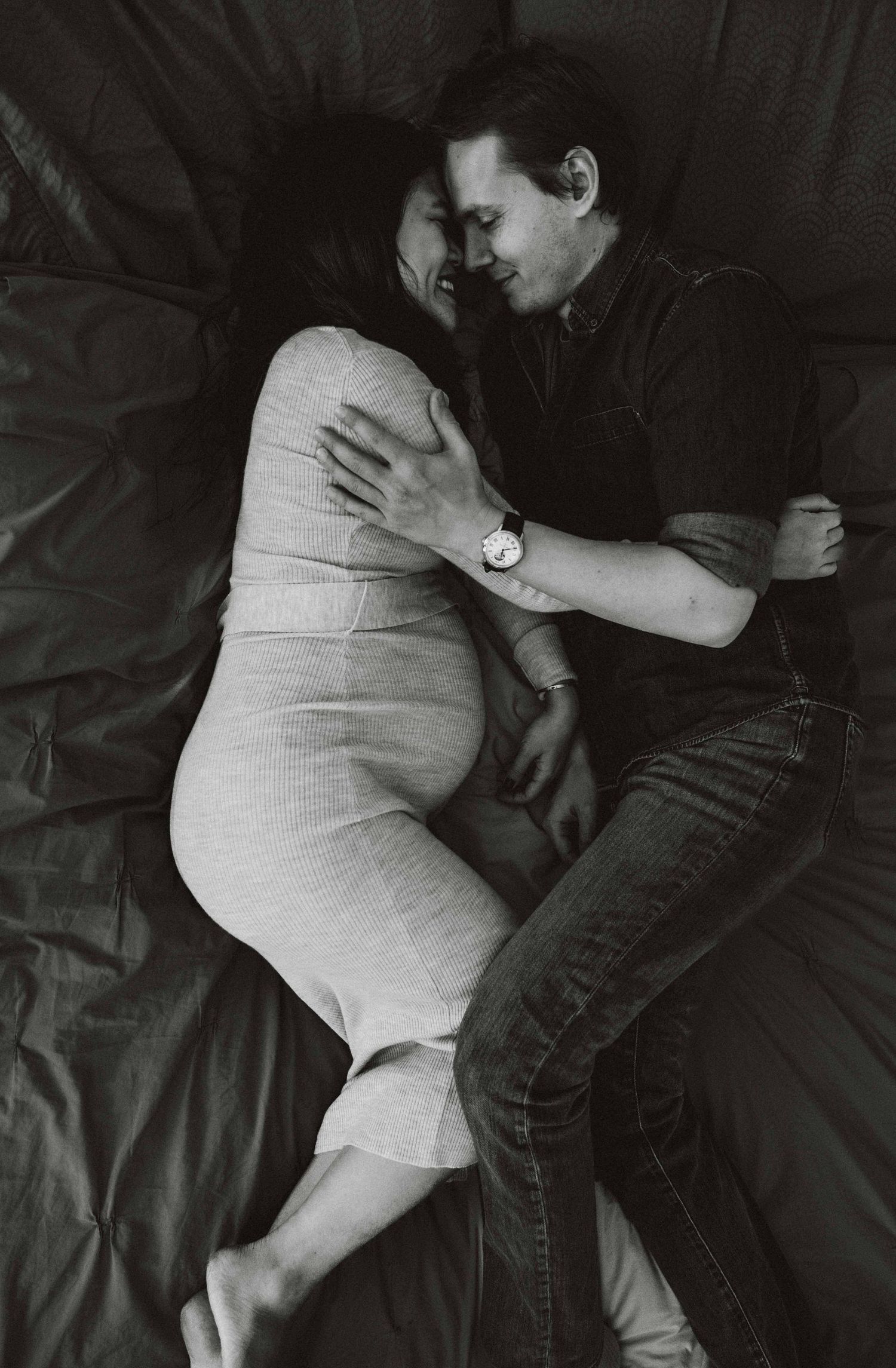 Black and white intimate maternity photos showing couple laying together on dark bedding.