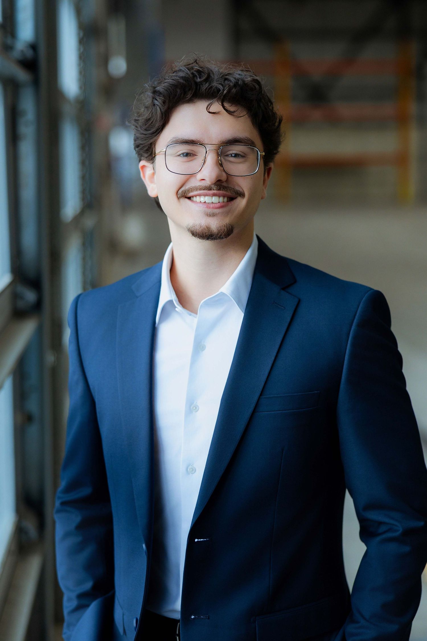 Young professional man in navy suit and glasses smiling confidently in modern office building.