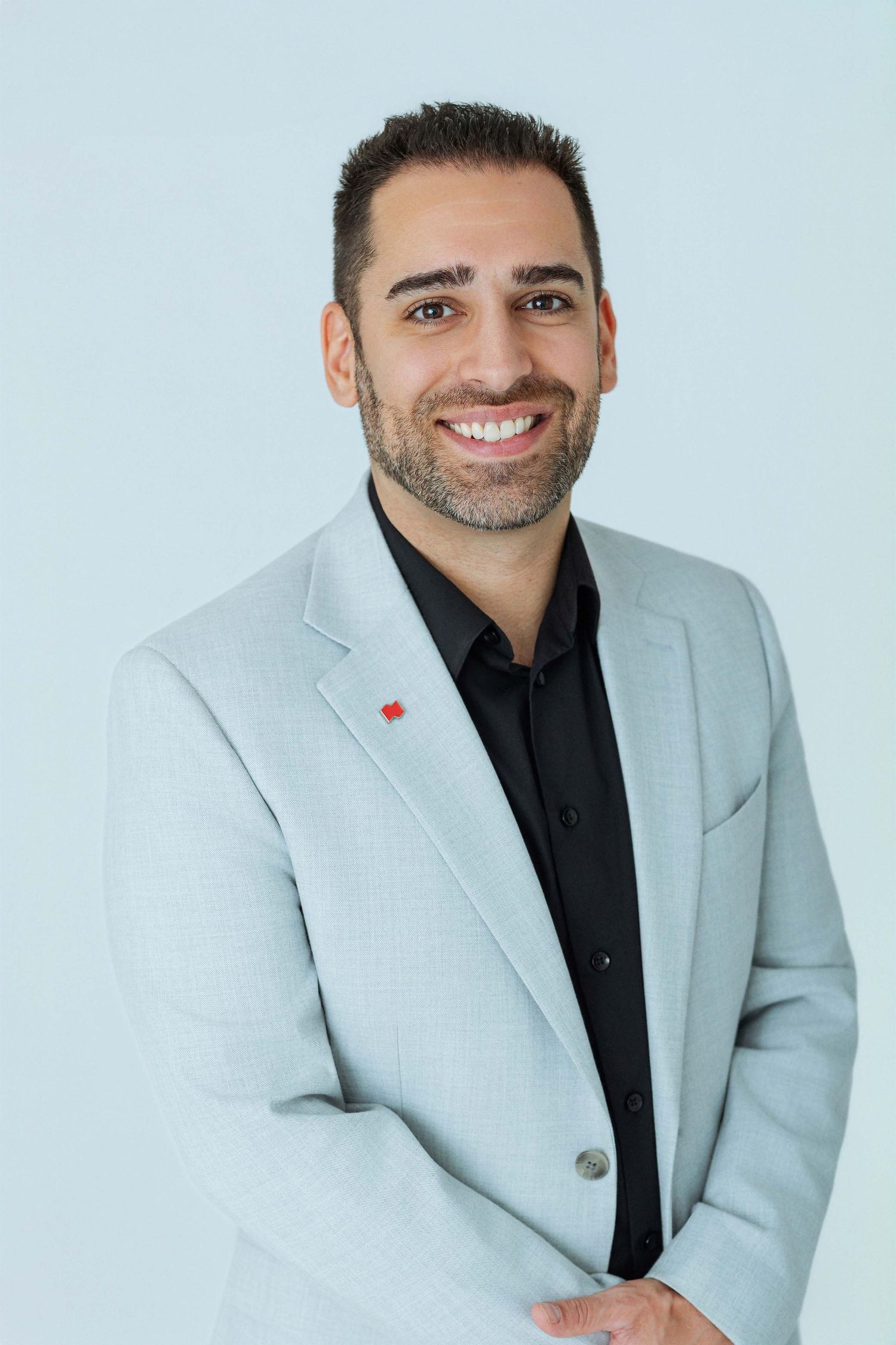 Smiling man in light gray blazer and black shirt poses confidently against a light blue background.