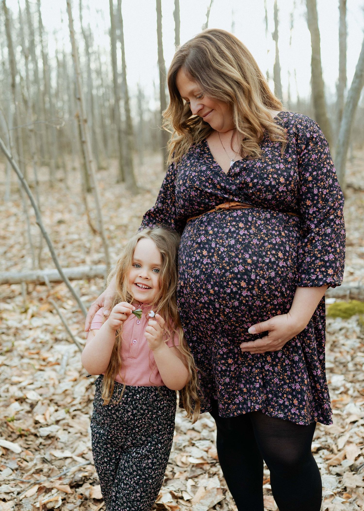 Pregnant woman in floral dress smiling at young daughter in autumn woodland setting.