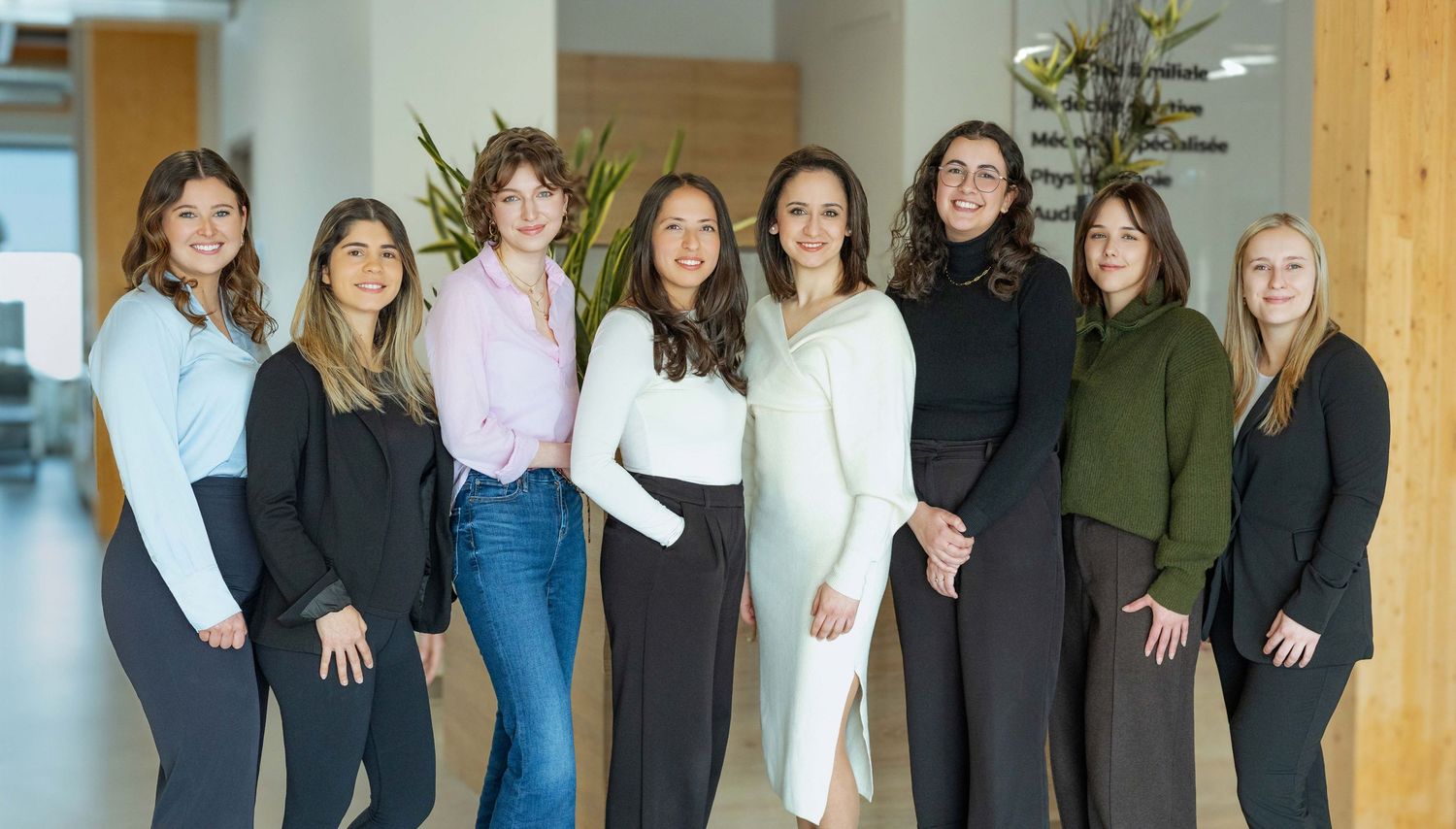 A group of people stand together smiling in a row against a light colored wall.