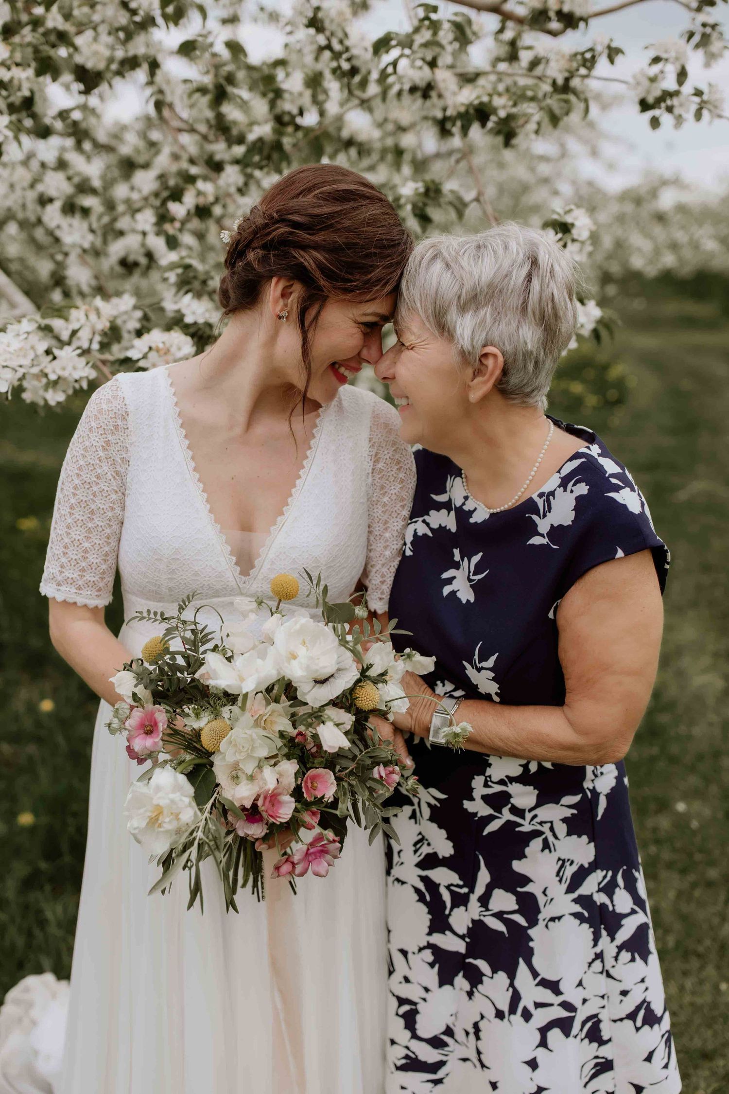 A tender moment between a bride in a lace dress and woman in floral print holding a beautiful bouquet.