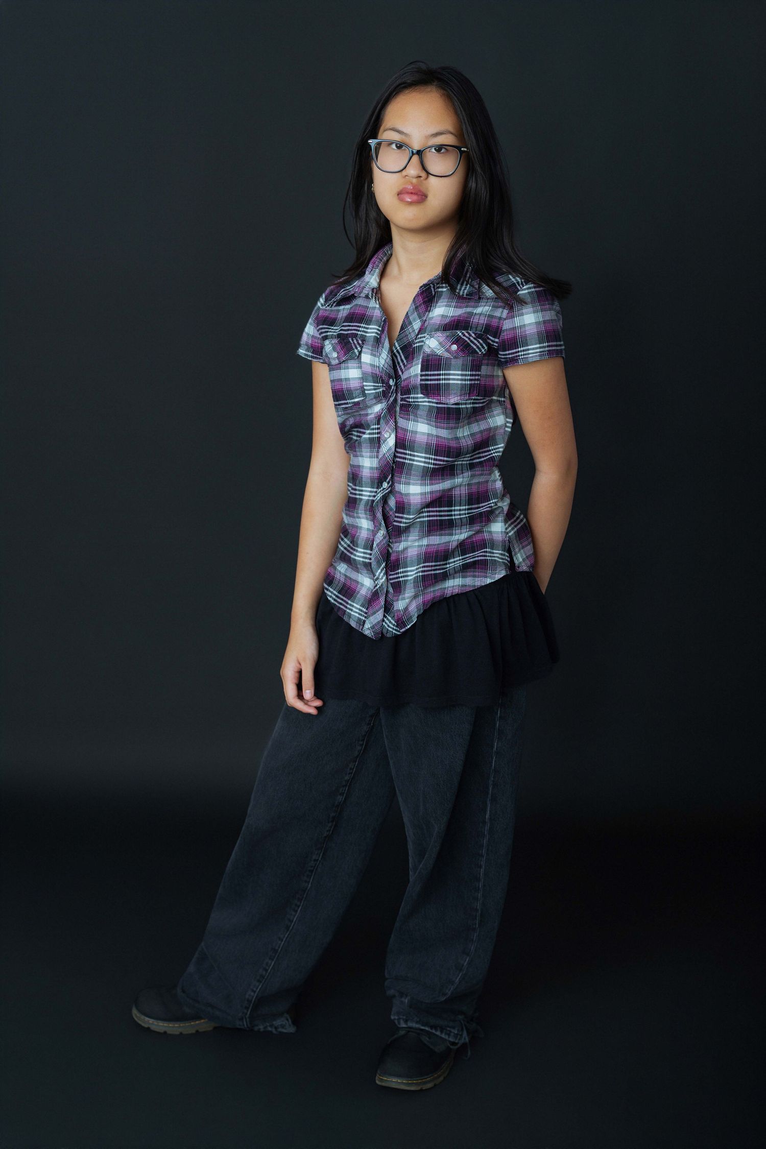 Young Asian girl in plaid shirt, glasses, and wide-leg jeans posing against dark background.