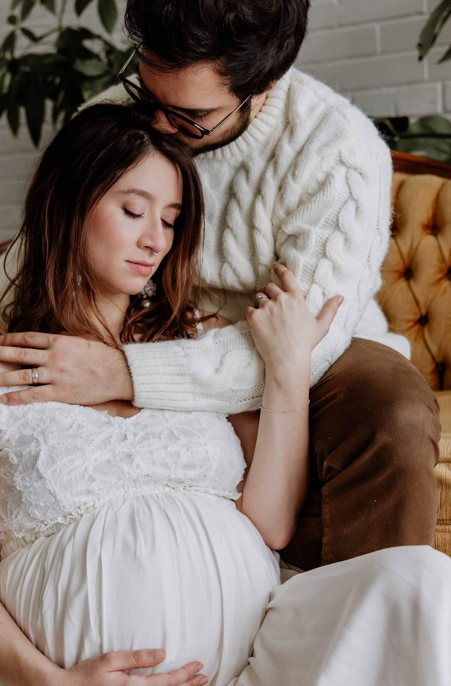 Intimate indoor maternity photo with couple in white sweaters on vintage furniture.