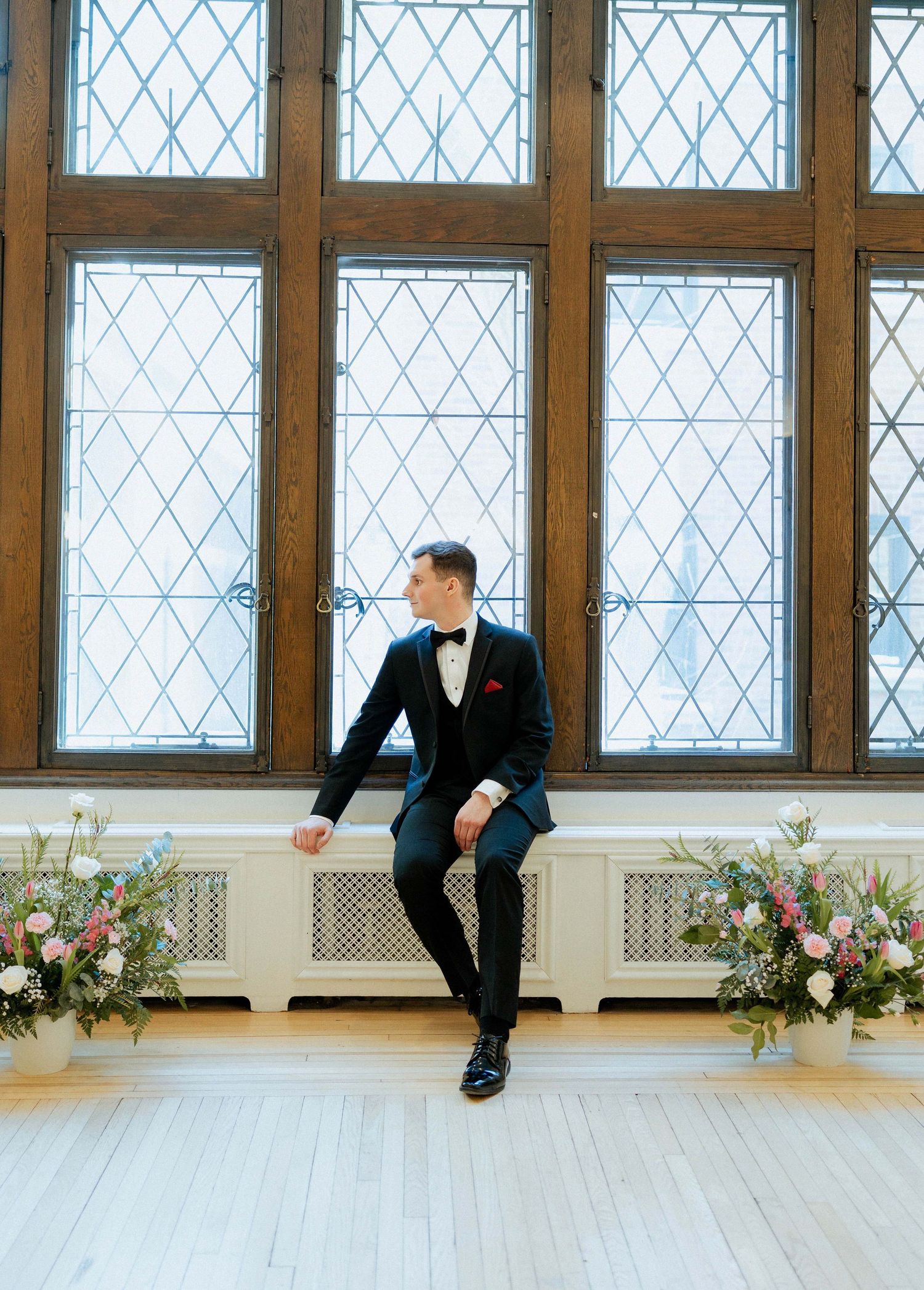 Elegant groom in black tuxedo sits by ornate window with diamond pattern glass panes and floral arrangements.