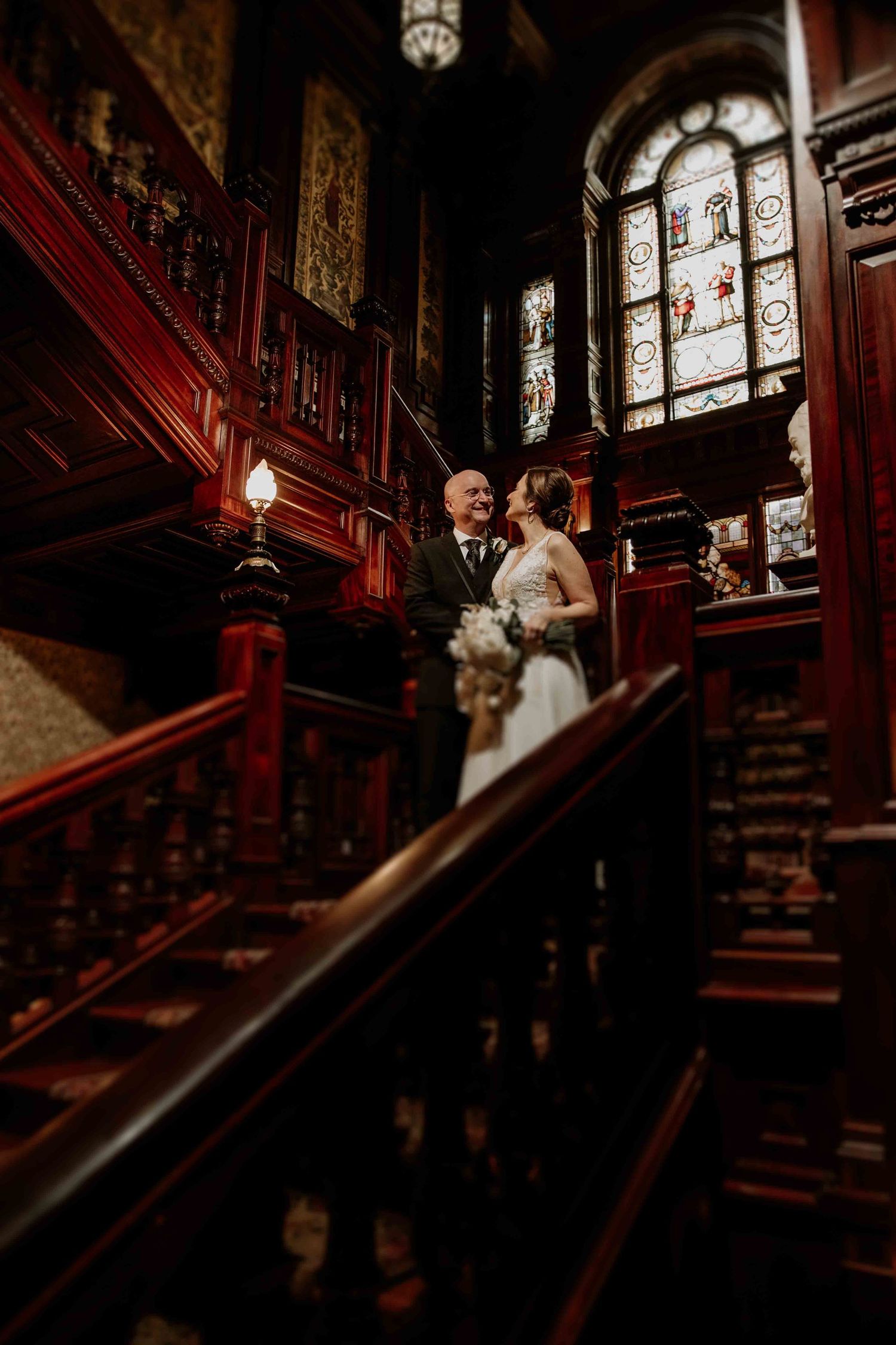 Dramatic staircase wedding photos in historic venue with stained glass windows and rich wooden details.