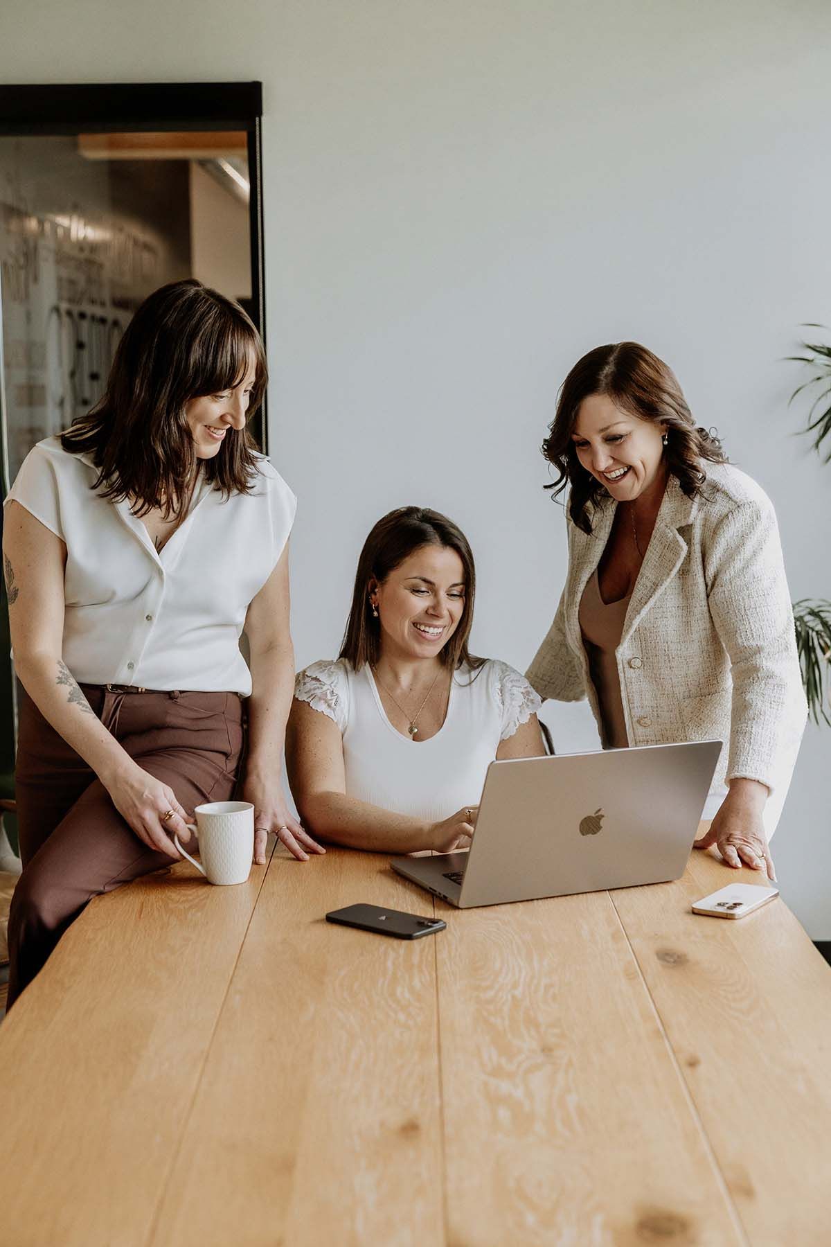 Three women smiling and collaborating around a wooden table with a MacBook laptop in a bright modern office.