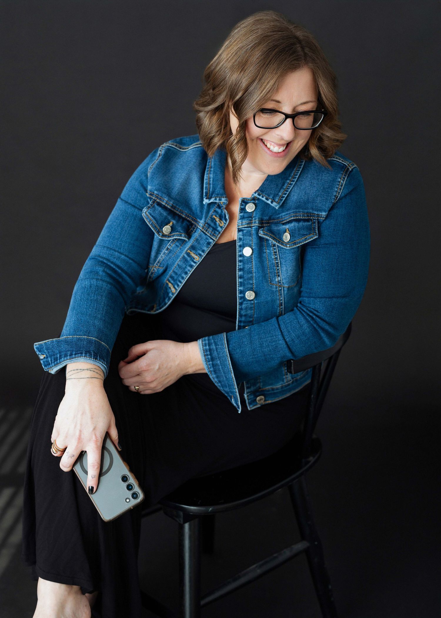 Woman in denim jacket and black dress sits on stool, smiling while holding a smartphone against dark background.