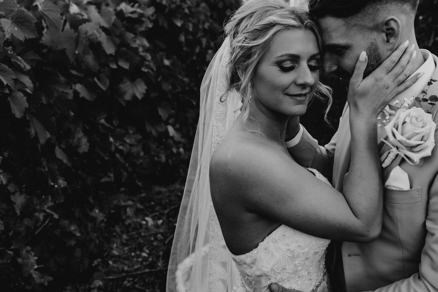 Romantic wedding couple embraces against stone wall backdrop in black and white photography.
