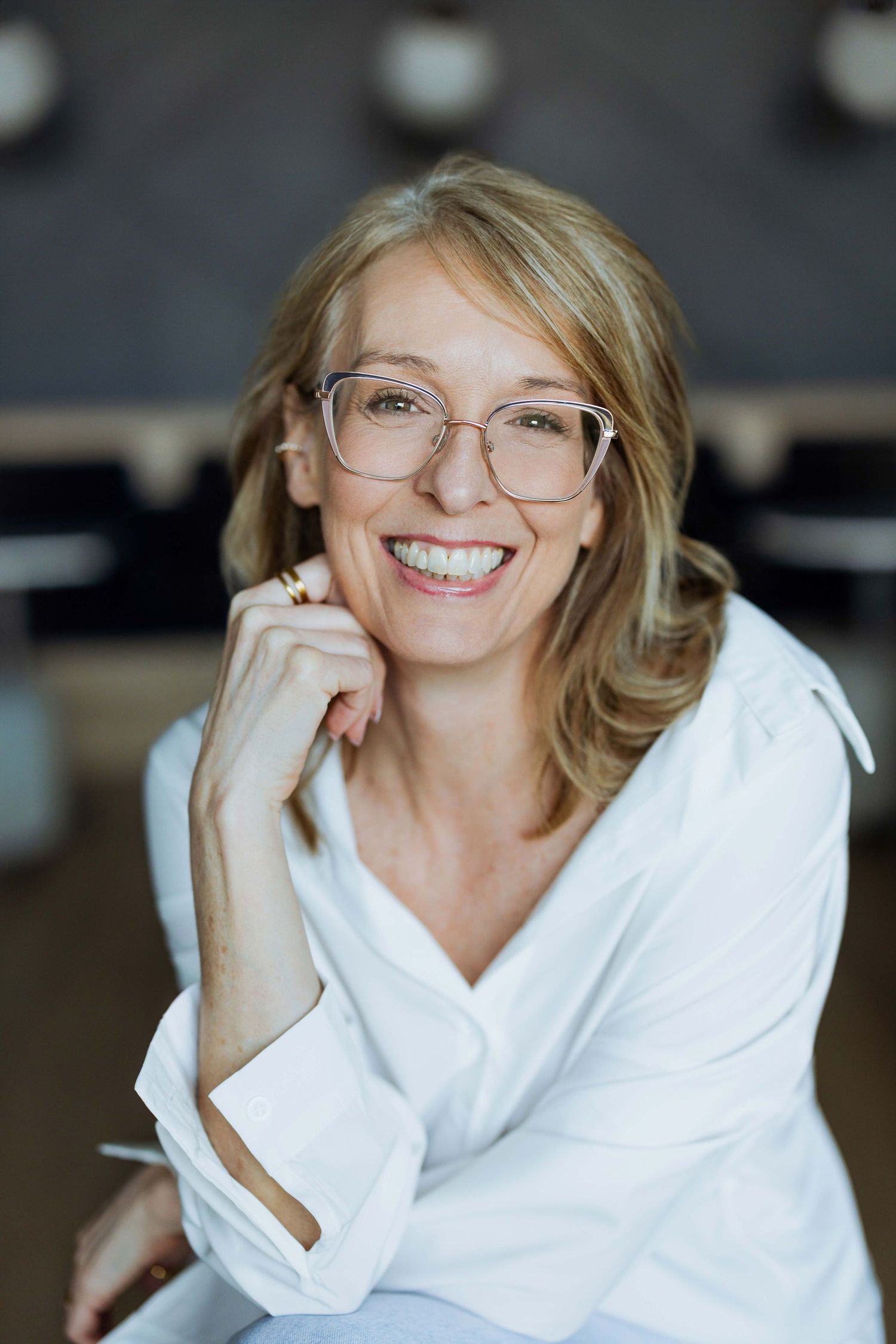 Smiling middle-aged woman with blonde hair and clear glasses wearing a white shirt, posing confidently indoors.