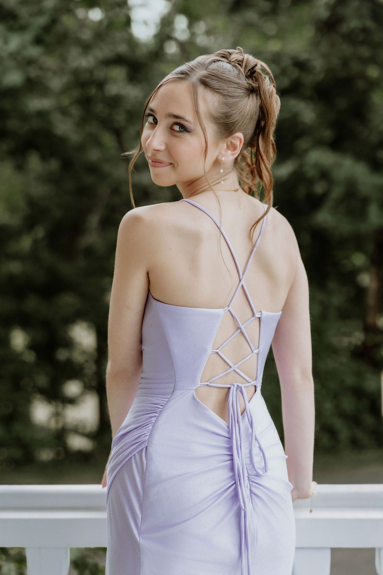 Young woman in lavender lace-up back formal gown posing outdoors, looking over shoulder with elegant updo hairstyle.
