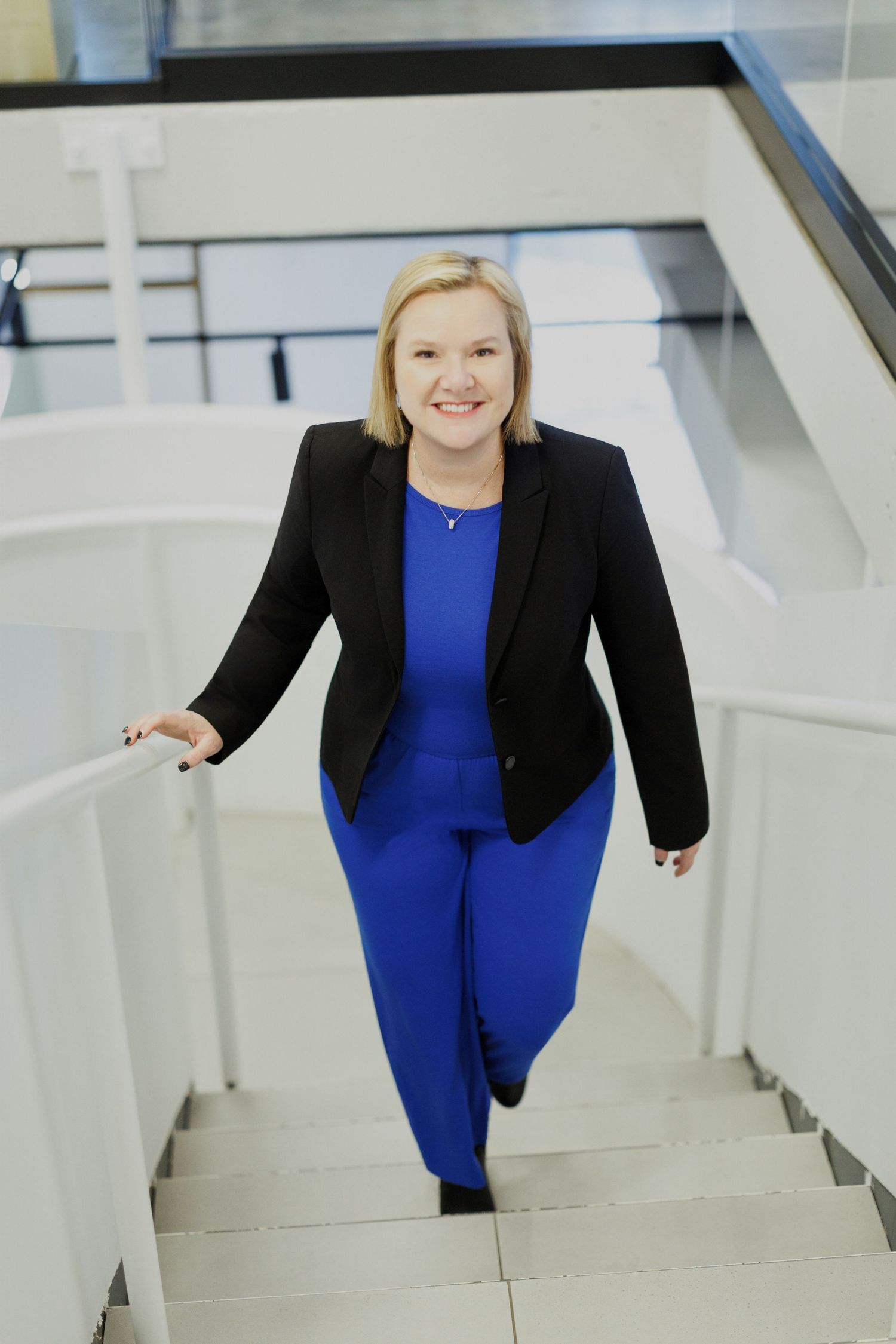 A person wearing a royal blue dress and black blazer poses on modern white stairs in a professional setting.