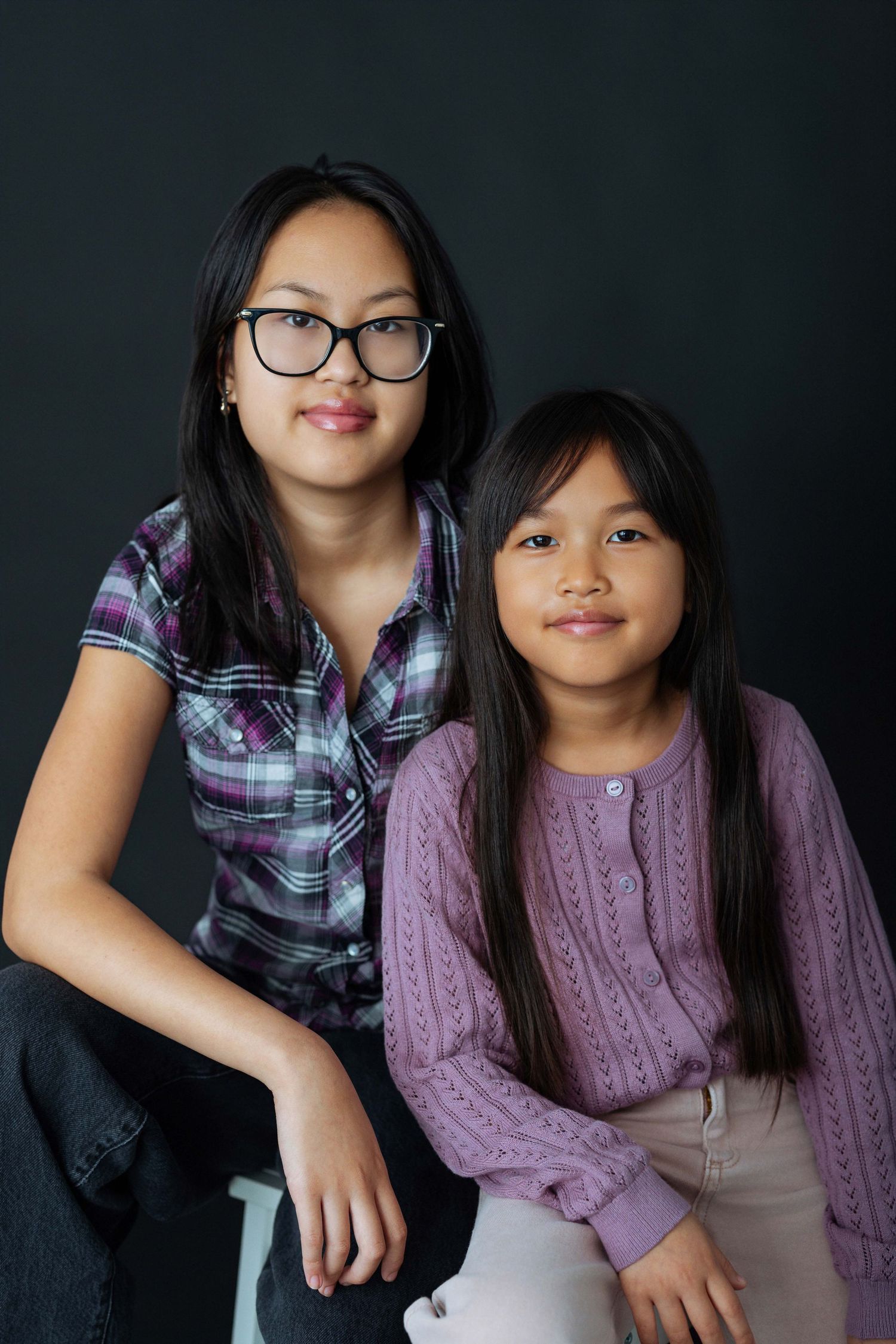Two Asian girls, older wearing glasses and plaid shirt, younger in purple sweater, posing together against dark background.