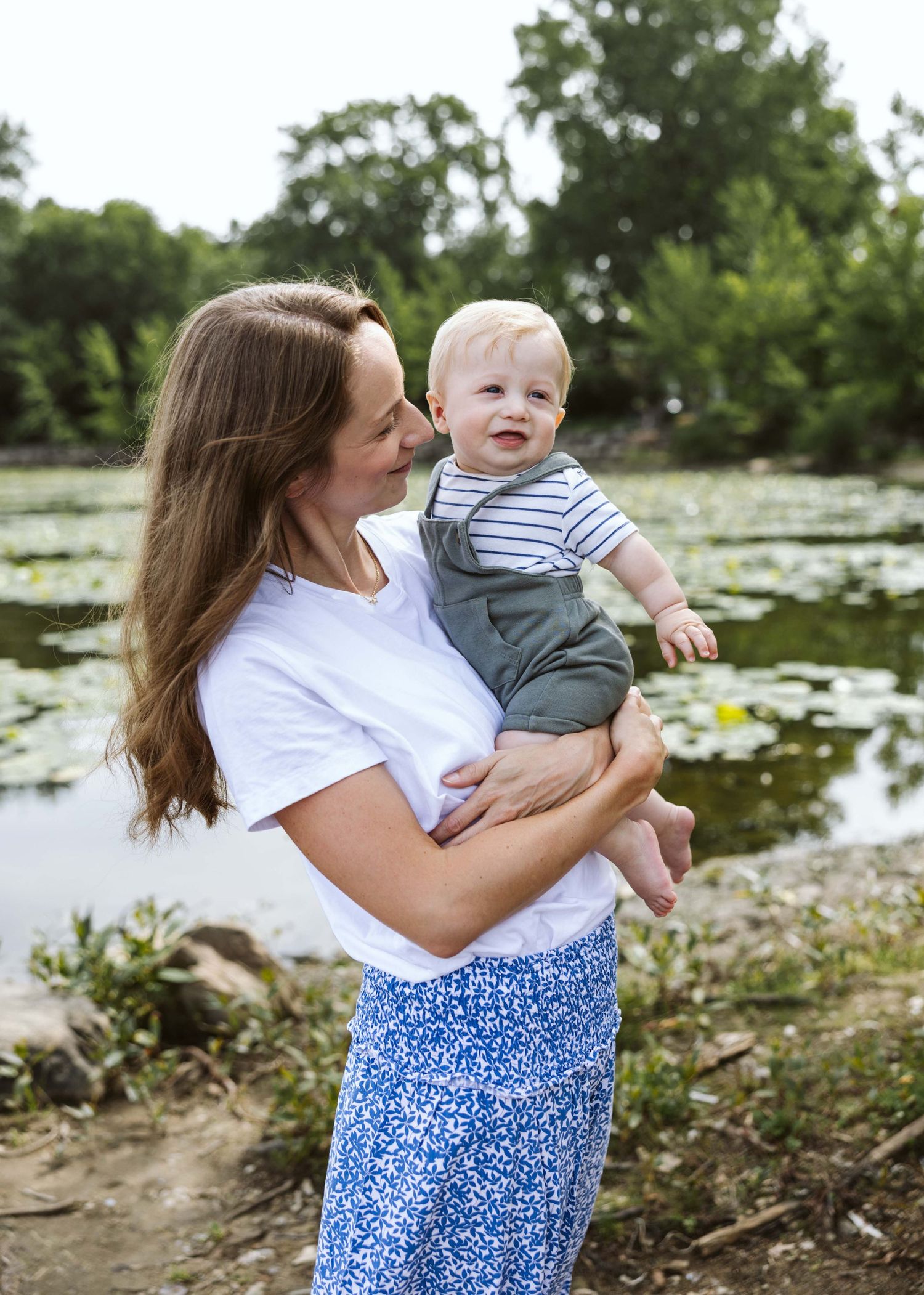 A smiling mother holds her happy baby boy near a serene lily pad pond surrounded by lush green trees.