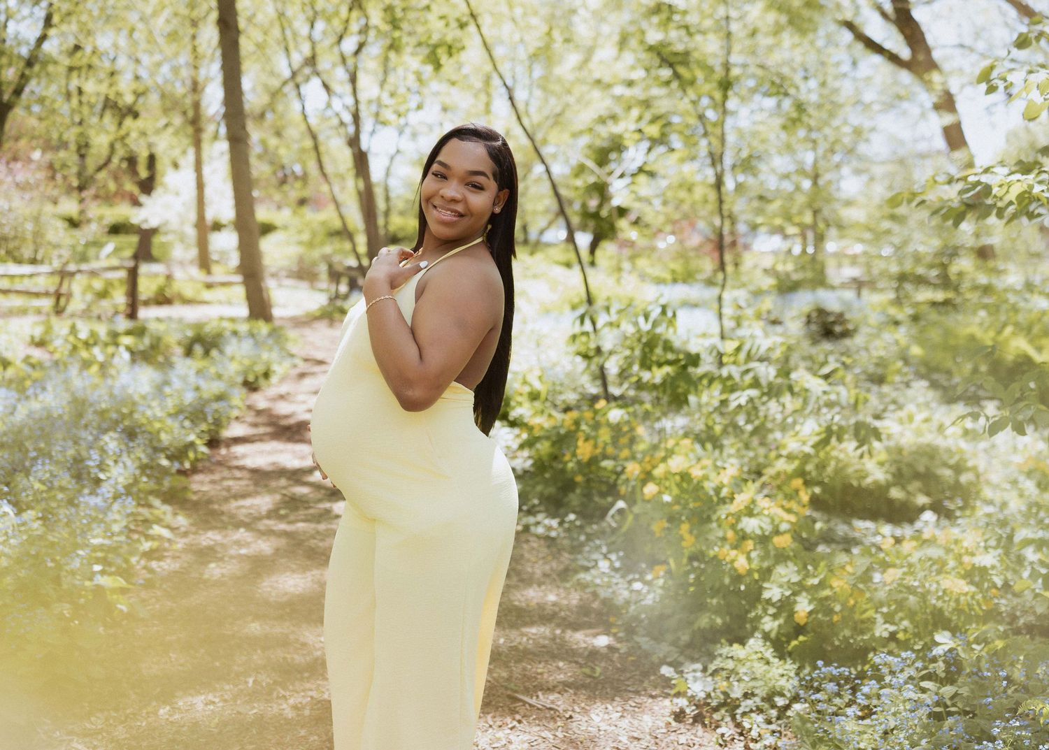 Pregnant woman in yellow dress posing outdoors in a lush green garden during a maternity photoshoot.