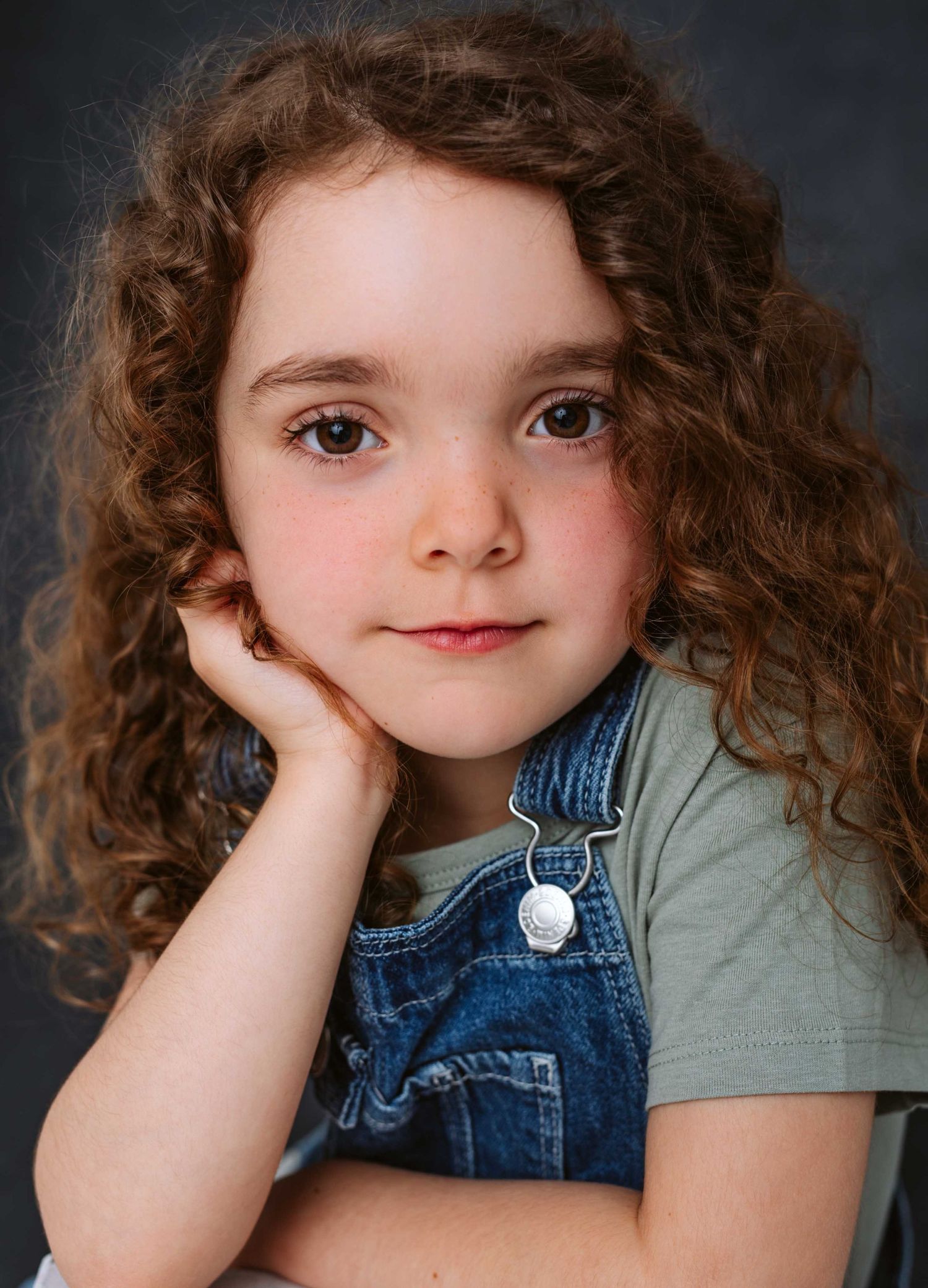 Child in denim overalls and green shirt poses with hand on cheek against dark background. , indéfendable, Casting agence peanut