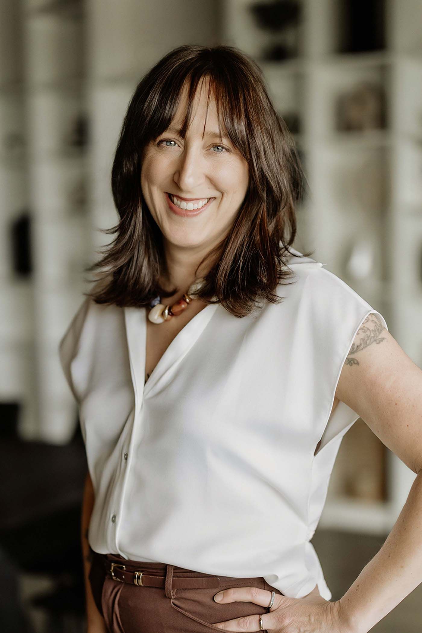 Smiling brunette woman with bangs in white sleeveless blouse and brown pants poses with hand on hip.
