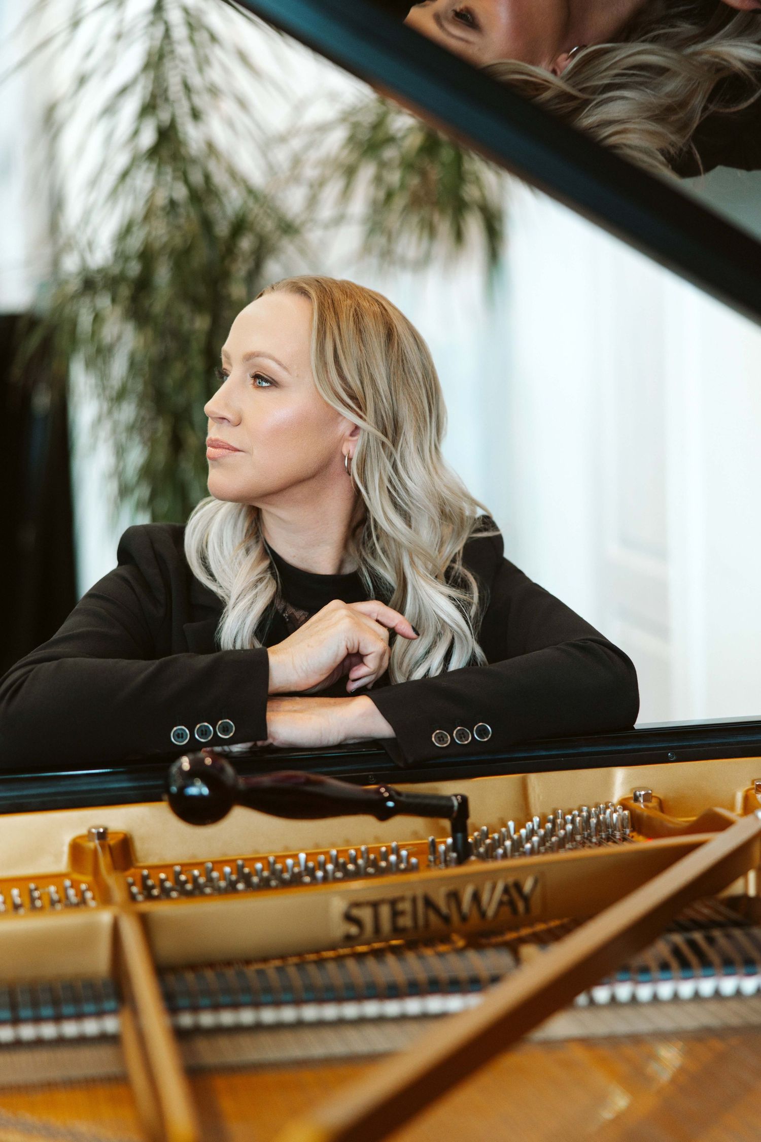 Blonde woman in black jacket resting arms on open Steinway grand piano, gazing thoughtfully to the side.