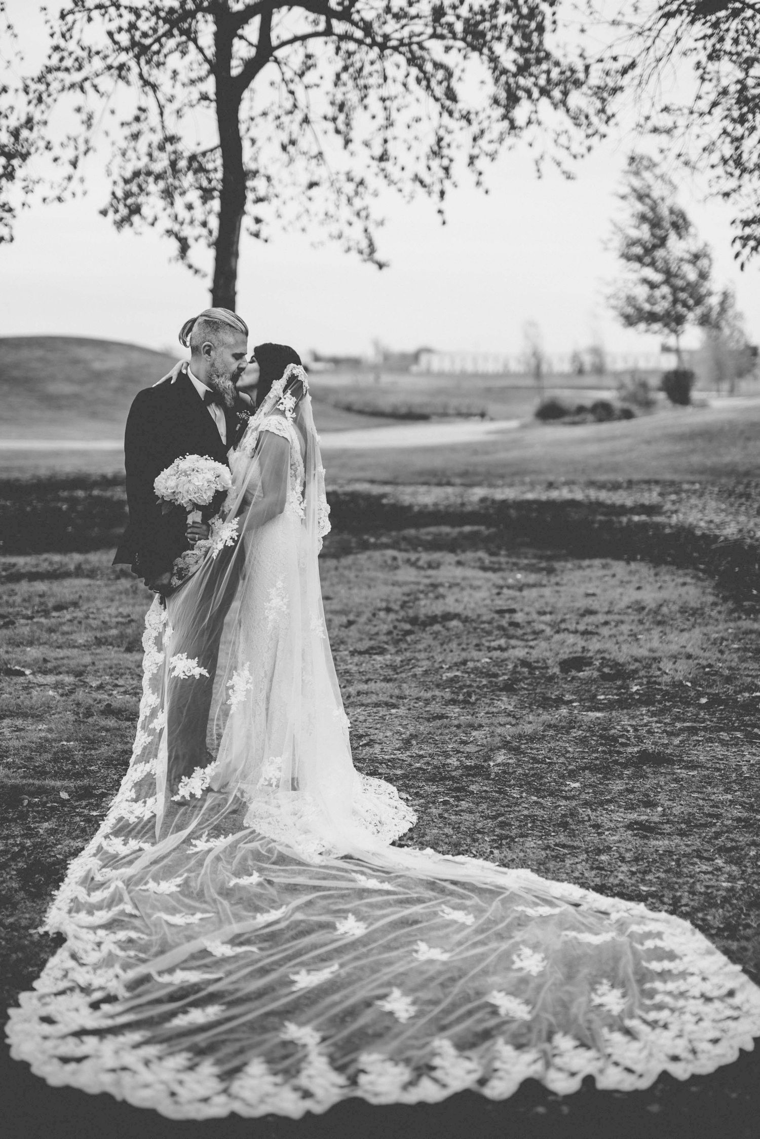 Wedding couple sits on grass with bride's elaborate lace train spread beautifully behind them.