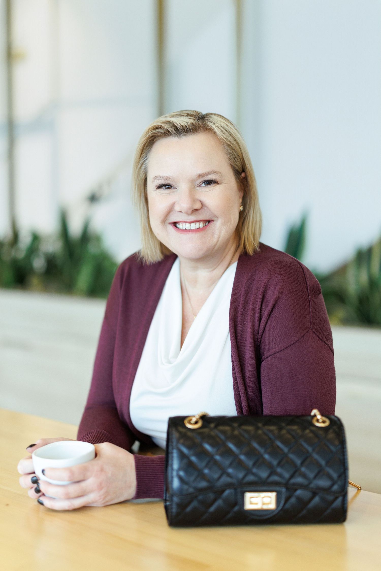 Professional portrait of someone in a burgundy cardigan and white top sitting at a table with coffee and a black quilted handbag.