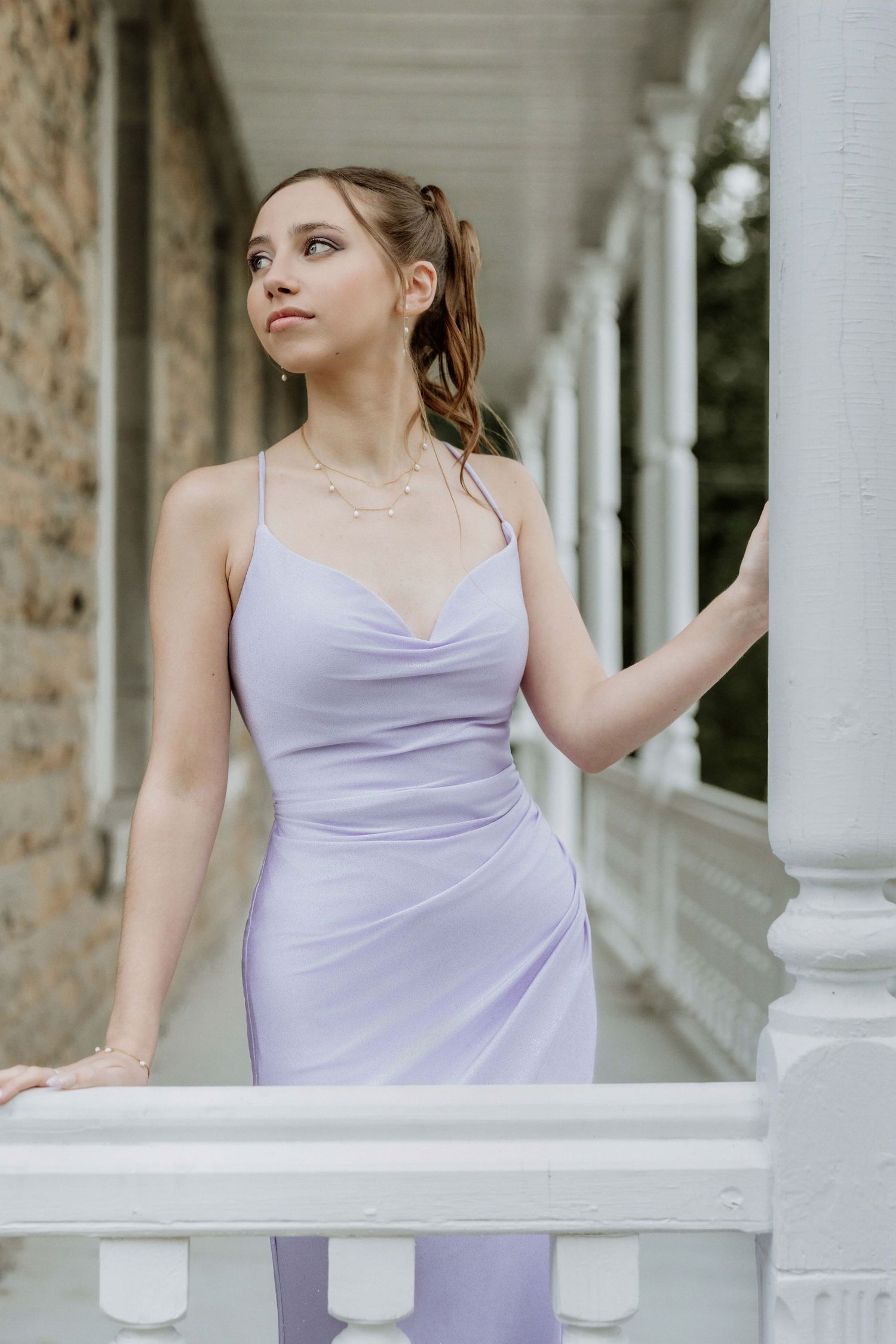 Young woman in lavender spaghetti strap dress poses gracefully by white porch railing of historic building.