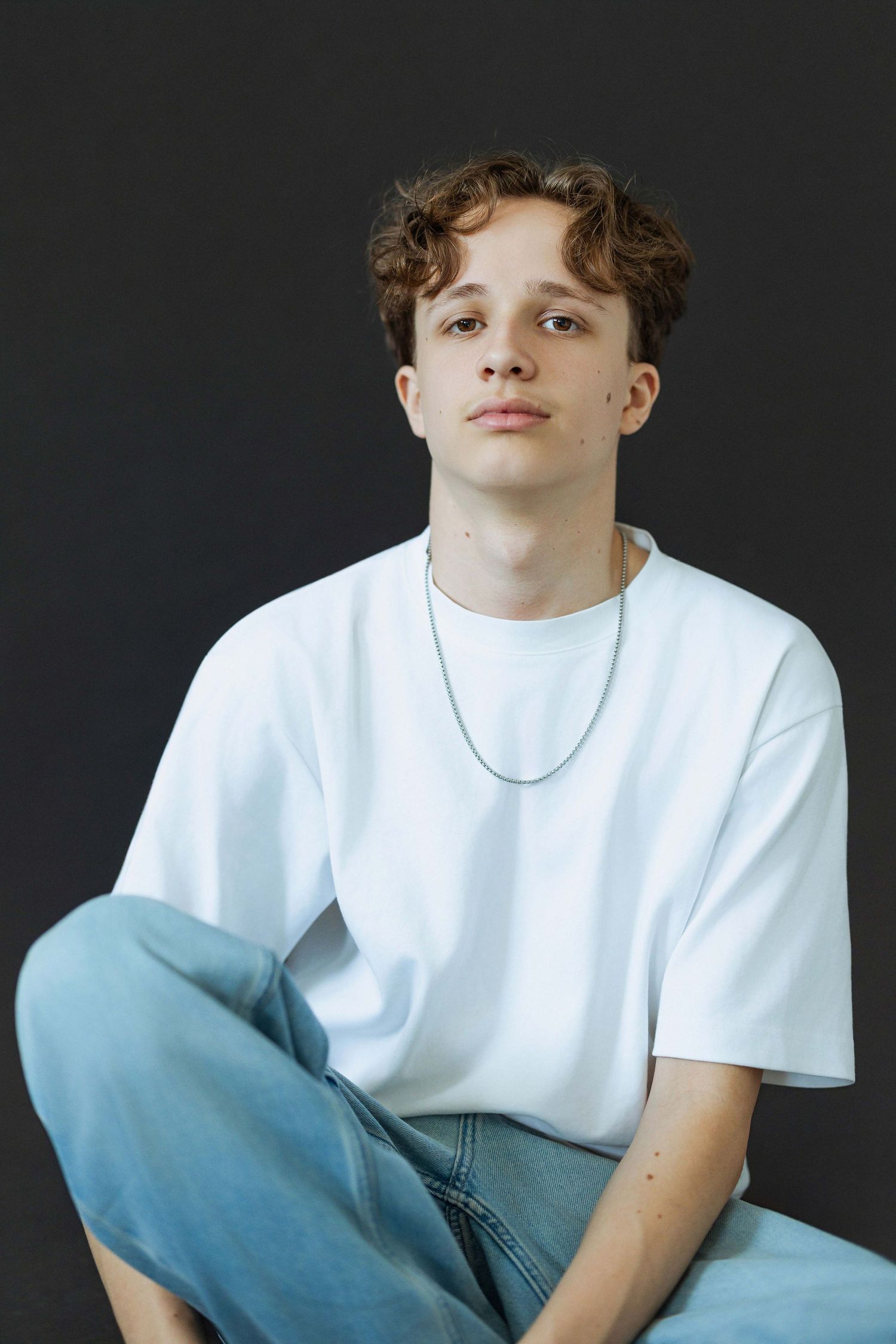 Young man with curly hair sitting cross-legged, wearing white t-shirt, jeans, and silver chain necklace against dark background.