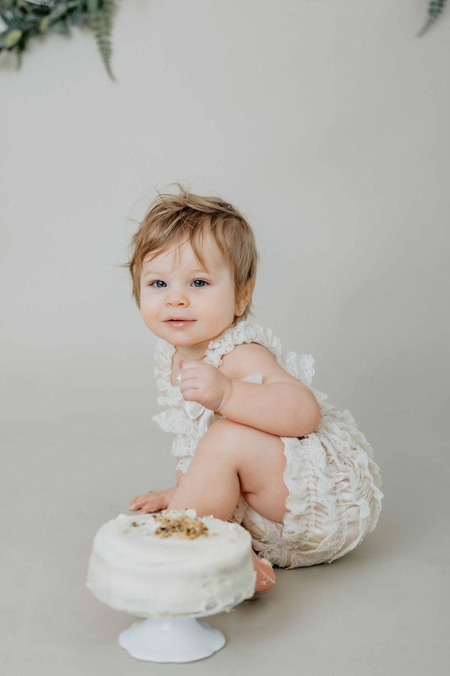 Baby in white lace outfit sitting next to cake on white pedestal.