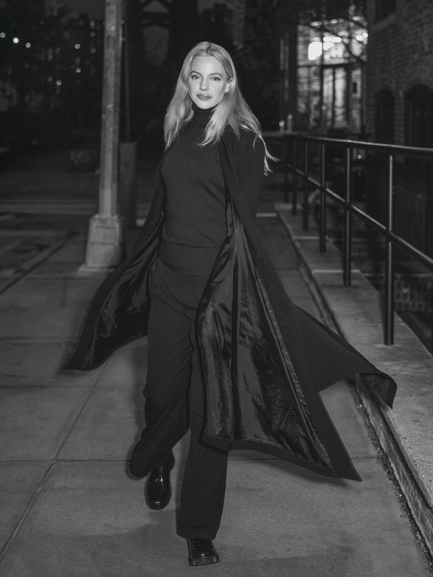 Black and white photo of a figure in flowing dark clothing and long hair walking dramatically along a railing at night.