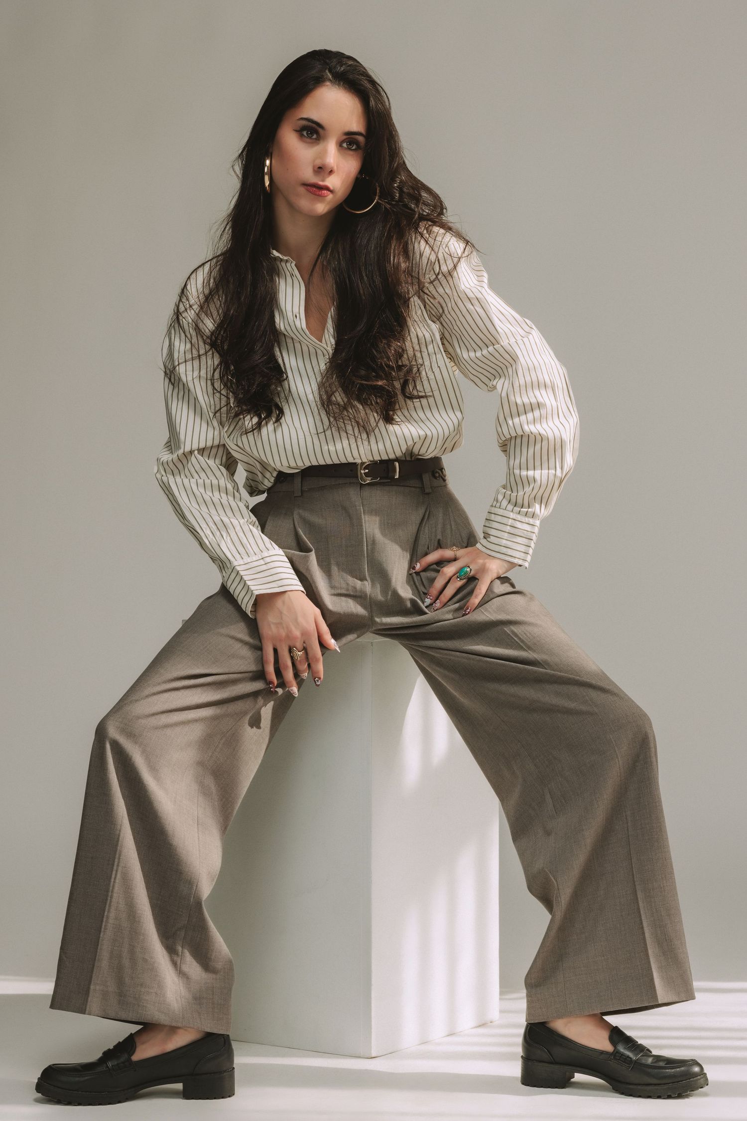 Woman in striped shirt and wide-leg trousers sits on white cube, wearing loafers and hoop earrings, studio photo.