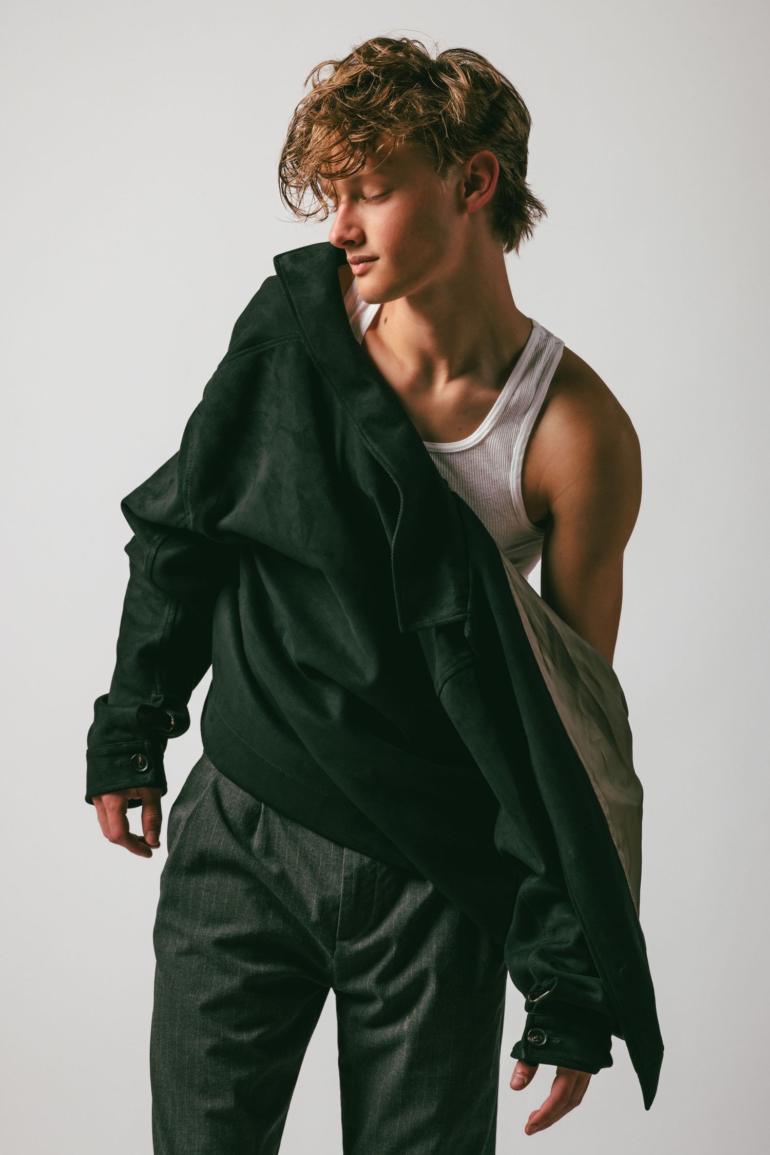 Young man in black oversized jacket, white tank top, and dark pants posing against a light gray background.