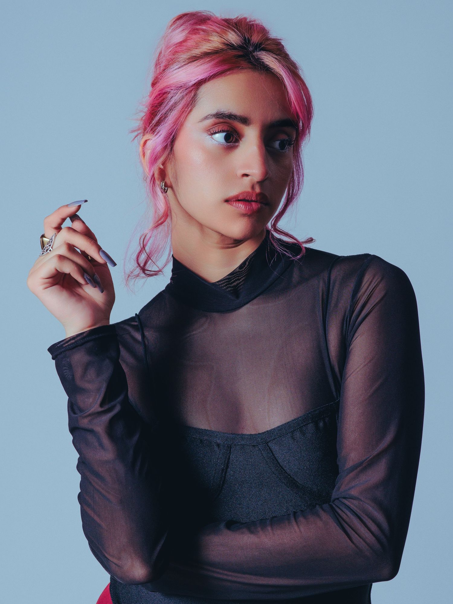 Person with pink hair wearing a sheer black long-sleeve top and dark bustier poses against light blue background.
